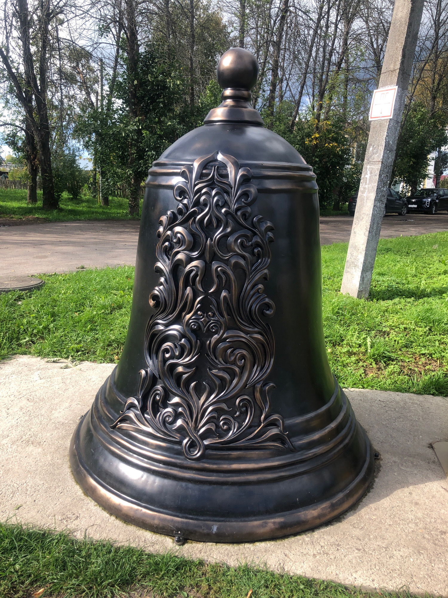 A large, dark bronze bell with intricate ornamental carvings is displayed outdoors, resting on a concrete base. The bell is positioned in the foreground, dominating the frame. It features a rounded top with a small finial and a flared base. The decorative elements on the bell are curvilinear and detailed, appearing to be cast metal with a lighter, possibly copper or bronze, patina.
Behind the bell, a grassy area leads to a paved road. Further back, a line of trees with sparse foliage suggests an autumn setting. A tall, rough concrete post stands to the right of the bell, leaning slightly. A small, square white sign with red border is affixed to this post, displaying a symbol that appears to be a red circle with a diagonal line through a silhouette of a person. Parked cars are visible on the road in the background. The sky is overcast with some patches of blue. The scene is set in the city of Tutaev, Russia, near a building with a white facade and a red roof, possibly a church or administrative building.