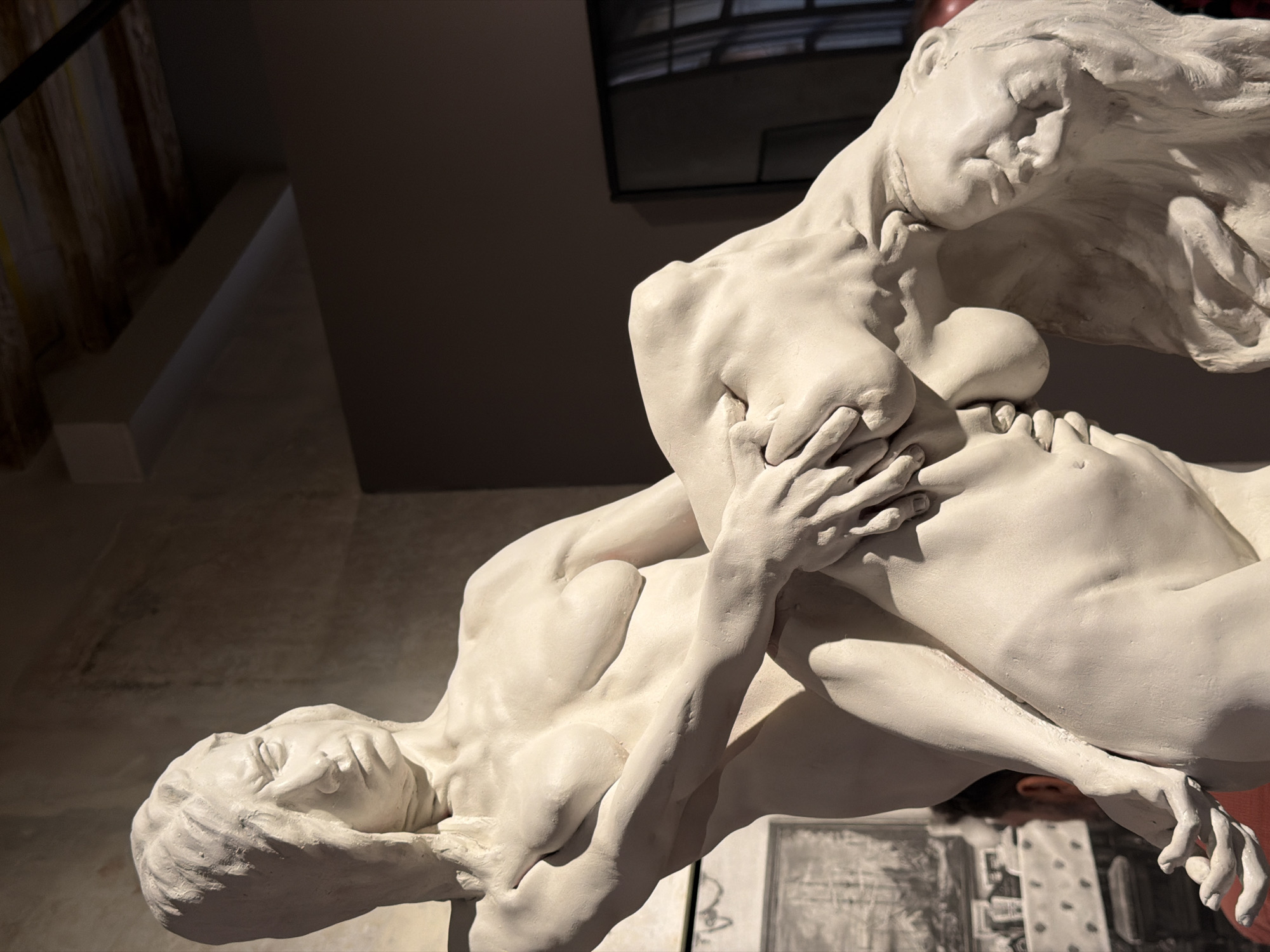 The media file is a close-up, low-angle shot of a white, plaster or clay sculpture depicting two intertwined figures. The figures are sculpted in a dynamic, almost fluid pose, with one figure appearing to be on top of or embracing the other. The sculpture is rendered with a high degree of anatomical detail, emphasizing musculature and form.
The upper figure's head is tilted back, with long, flowing hair depicted as if caught in motion. Their eyes are closed, and their facial expression suggests a moment of intense emotion, perhaps ecstasy or surrender. Their hands are tightly clasped around the torso of the lower figure. The lower figure's head is angled downwards, with their face obscured and their body contorted beneath the embrace of the upper figure.
The sculpture is illuminated by what appears to be artificial lighting, casting strong shadows that accentuate the contours and musculature of the figures. The background is dimly lit and out of focus, but hints of a dark, textured wall and possibly some vertical elements like curtains or drapery can be seen. In the lower portion of the image, a blurred background reveals what might be a display area in a gallery or museum, with a partial view of a framed artwork, possibly a print or drawing, featuring architectural elements.
The overall impression is one of passionate connection and dynamic movement captured in a static medium. The lighting and composition create a dramatic and intimate feel. Given the location context of Barcelona, Spain, this sculpture might be part of a museum collection or a public art installation. There is no visible text in the media file.