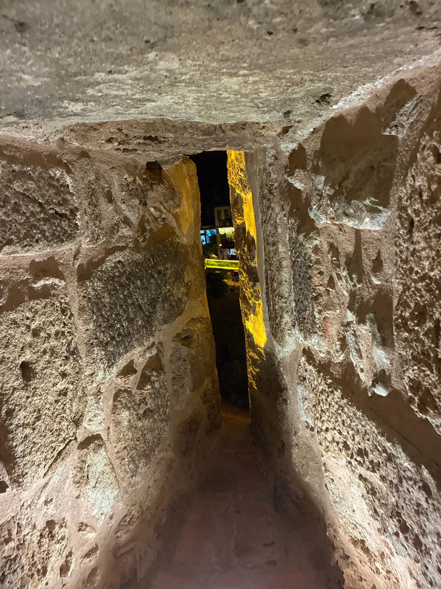 The image depicts a narrow, arched opening within a thick stone wall, likely a passage or window in the Alanya Castle complex in Alanya, Turkey. The rough-hewn stones of the wall are illuminated by artificial light, casting shadows and highlighting their textured surfaces. Through the opening, a dark passage leads to a brightly lit exterior scene. In the distance, illuminated by yellow lights, are glimpses of buildings with dark windows and balconies, suggesting an urban setting outside the castle walls. The ground within the passage appears to be made of reddish earth or paving stones. The overall impression is one of an ancient structure looking out onto a modern or older town.