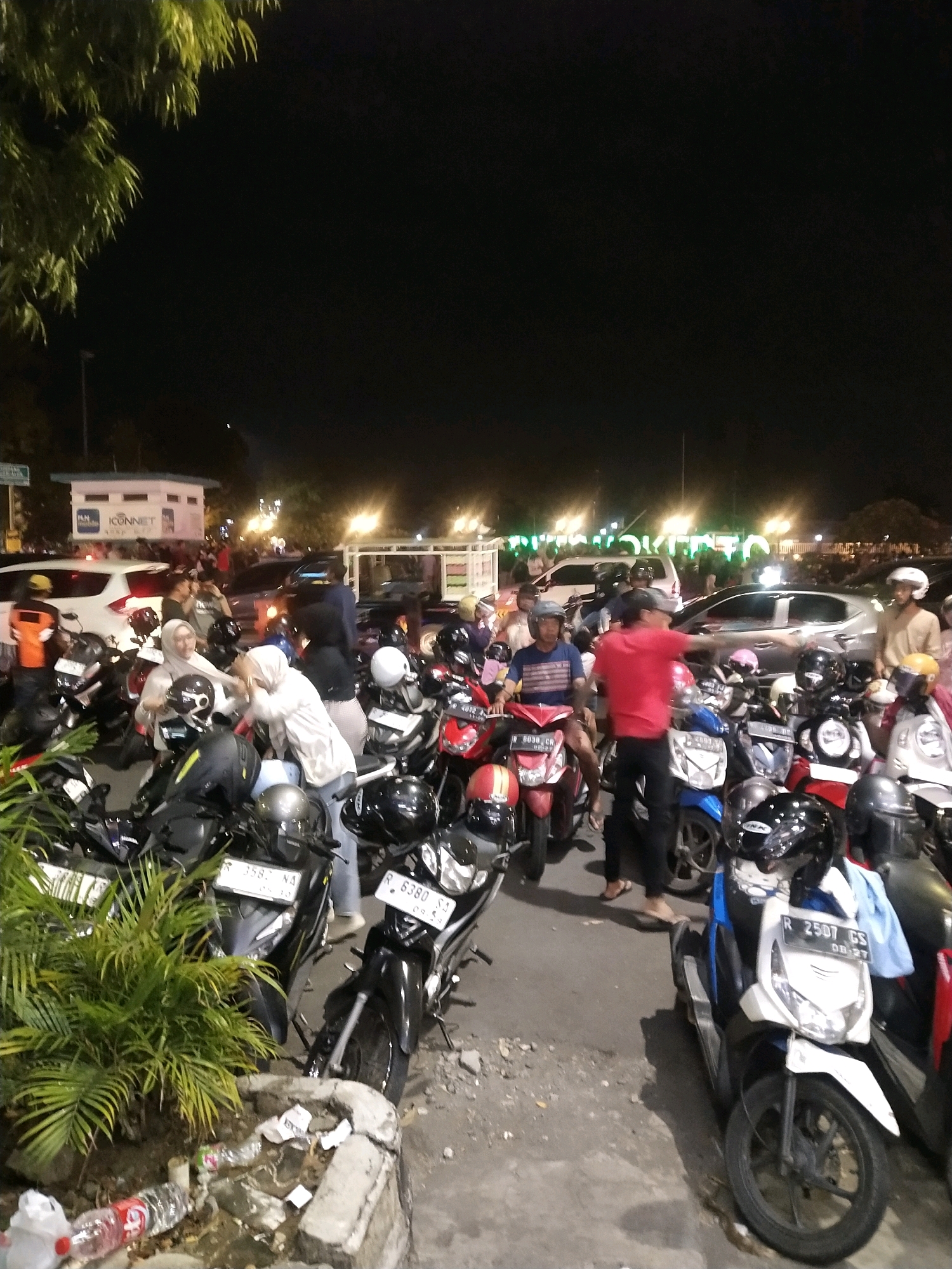 The image depicts a nighttime scene in a parking lot in Purwokerto, Indonesia. Numerous motorcycles are parked, with some people milling about. The lighting suggests the presence of streetlights and building illumination. In the foreground, a green plant is visible, and there is some litter on the ground. Several individuals are interacting, with some appearing to be preparing to leave or having just arrived. The overall atmosphere is that of a busy public space at night.
