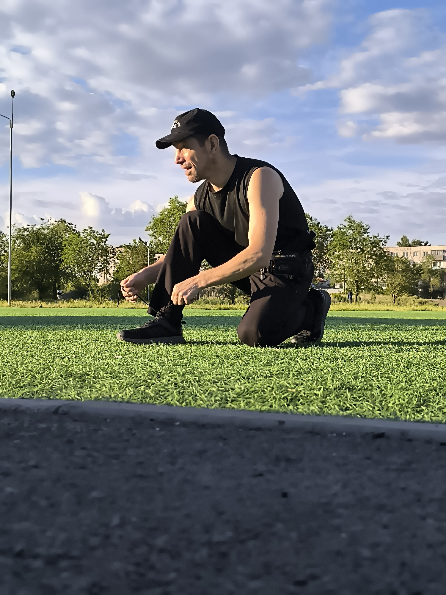 A man wearing a black baseball cap and a black sleeveless shirt is kneeling on a bright green, artificial turf field. He is in the process of tying his black athletic shoes. His dark pants are pulled up slightly, revealing his lower legs and ankles. The man is looking down at his shoes, and his facial expression suggests concentration.
The background of the image shows a clear blue sky with scattered white clouds. A tall, thin lamppost stands to the left of the man. Beyond the field, there is a line of green trees, and further in the distance, several apartment buildings can be seen. The edge of a dark, asphalt-like surface is visible in the foreground, blurred due to its proximity to the camera. The overall scene appears to be an outdoor sports facility, likely in a city park or recreational area.