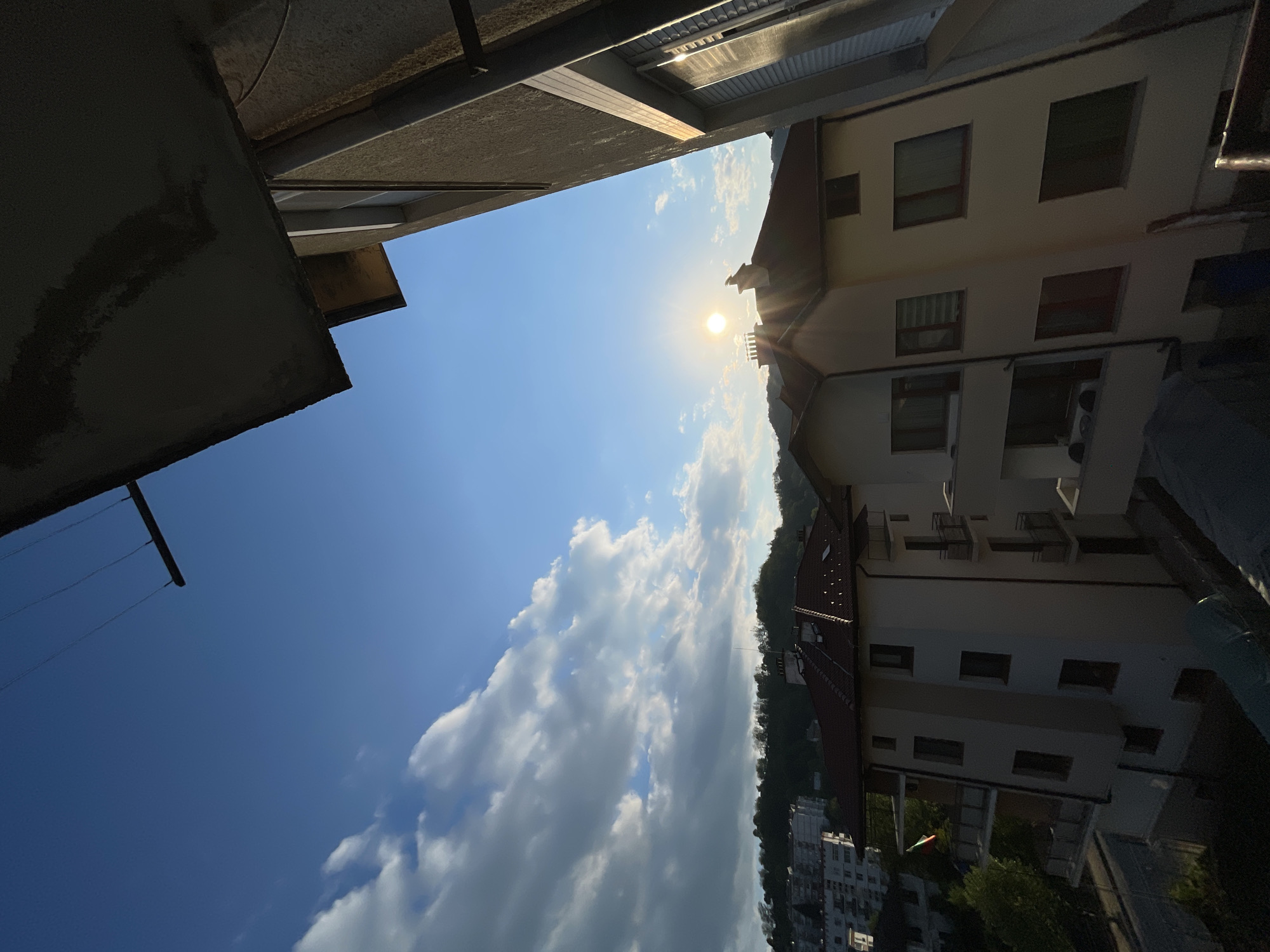 The image presents a view of the sky from between buildings. The sky is mostly blue with scattered white clouds and the sun is positioned above a hill with trees. Buildings are visible on either side of the frame, with architectural details such as windows, balconies, and rooflines. The photo was probably taken in Zlatograd, Bulgaria.