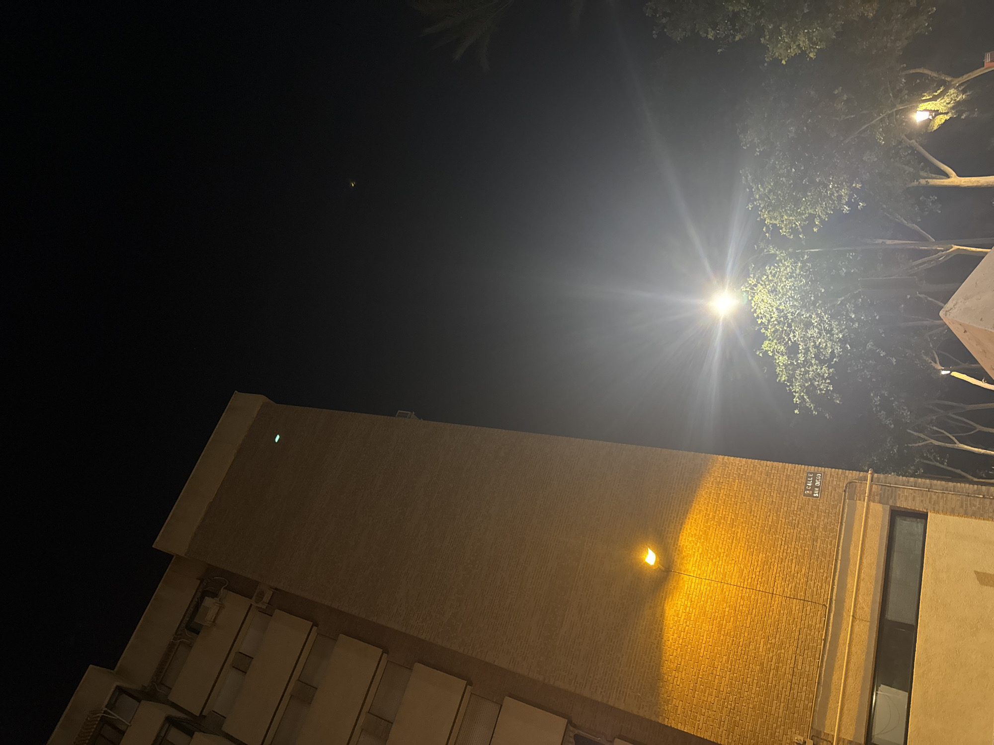 The image captures a nighttime street scene in Murcia, Spain. On the left, a tall brick building is illuminated by a warm yellow streetlamp, casting a glow on the textured facade. A sign reading "2 CALLE SAN DIEGO" is visible on the brick wall. In the background, trees with lush green foliage are lit by a bright streetlamp, creating a halo effect. Another streetlamp is visible further back, and a hint of a palm tree is seen on the right. The sky is dark, with a faint haze, and two small, distant lights are discernible. The scene appears to be from a residential or urban area.