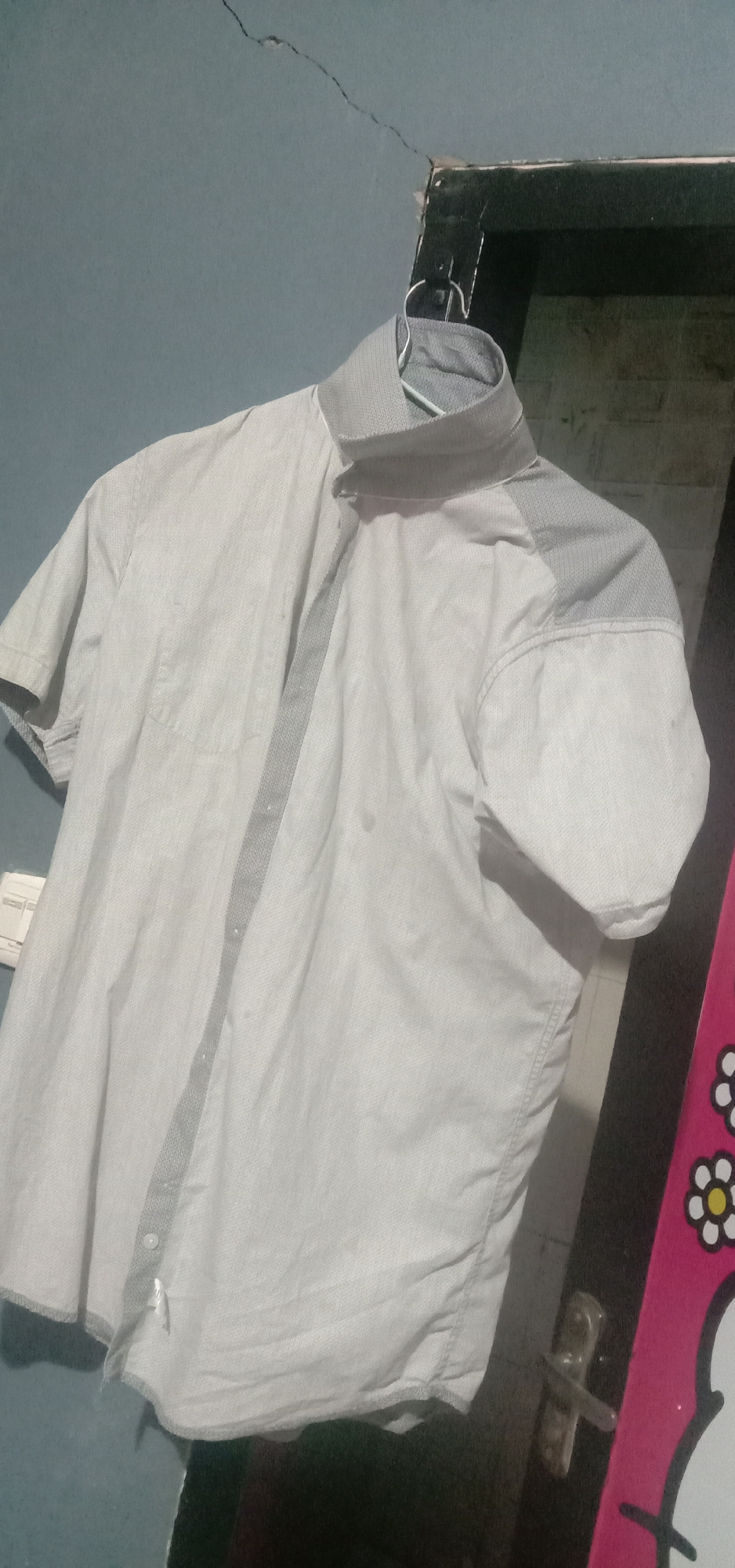 The image shows a light-colored, short-sleeved button-up shirt hanging on a hanger against a blue, cracked wall. The shirt has a patterned collar and a textured stripe down the front. There is a white light switch on the wall to the left of the shirt. To the right of the shirt, a dark doorway is visible, with a pink frame adorned with cartoon flower and character cutouts. The overall scene suggests an indoor domestic setting, possibly a bedroom or laundry area. The presence of a cracked wall could indicate an older building or possible structural issues. The lighting is dim, casting shadows on the shirt and surrounding objects. The image does not provide any specific clues to its location within the city of Kendari, Indonesia.