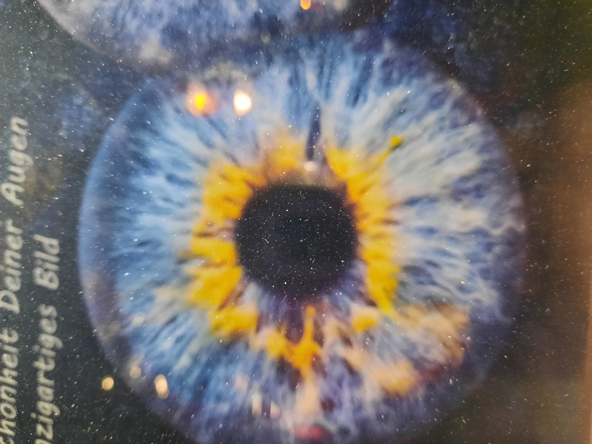 The image displays a close-up, unfocused view of a human iris with vibrant blue and yellow patterns, resembling a sunflower or a celestial body. The background is dark and speckled, suggesting a textured surface or dust. Text is visible at the top of the image, with the words "Schönheit Deiner Augen" (Beauty of Your Eyes) and "einzigartiges Bild" (unique picture) legible in a white font. The overall impression is abstract and artistic, with the iris being the central subject. There is no identifiable location in the city of Rosenheim, Germany, depicted in this image.