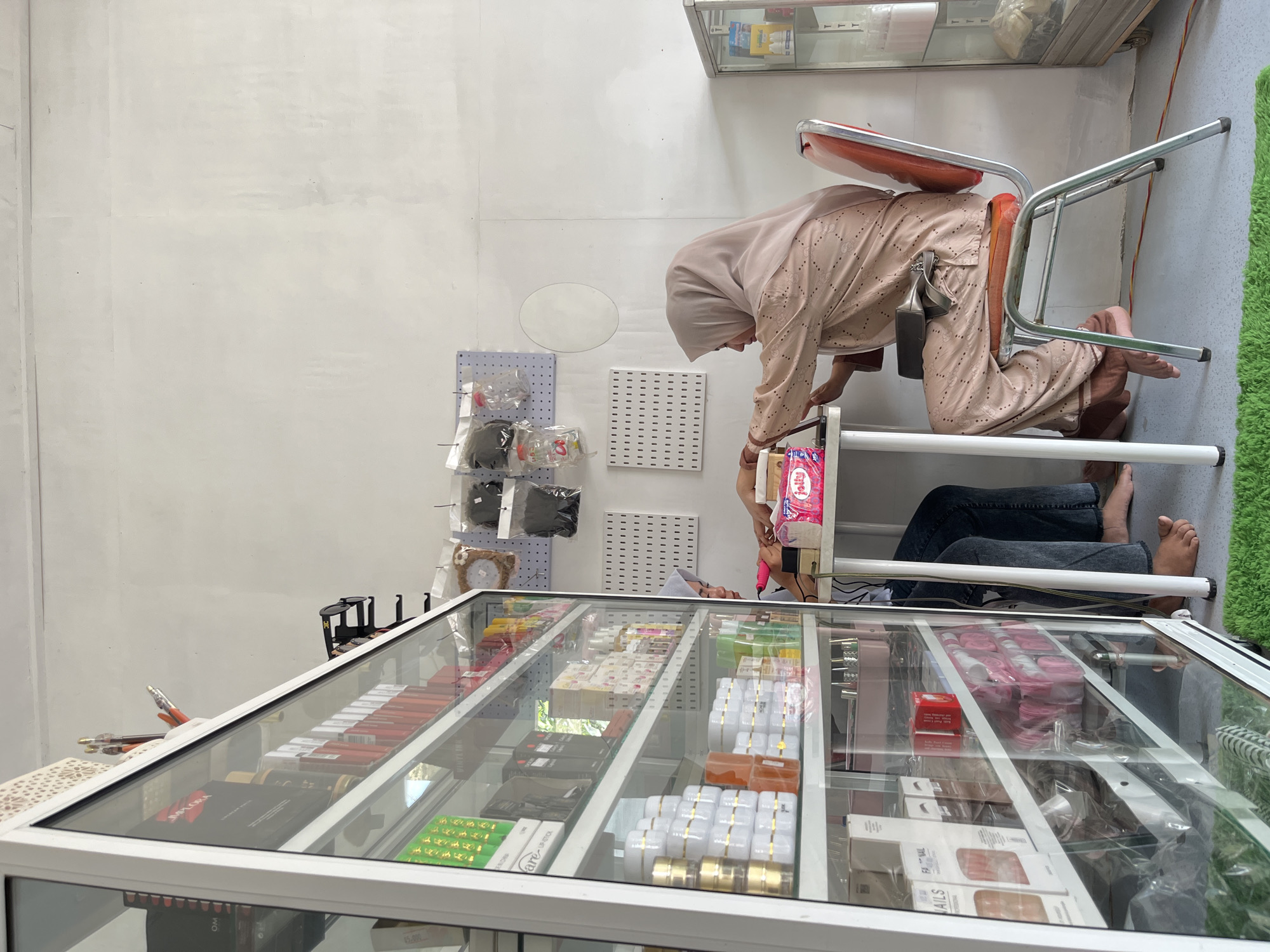 A rotated eye-level indoor shot shows a woman in a light tan hijab and patterned robe on an orange metal chair leaning over a glass display case, next to another person wearing jeans. The location appears to be Lhokseumawe, Indonesia, given the items displayed in the case, which include cosmetics and beauty products. The case is white-framed and contains a variety of items, including lipstick, skin creams, and various packages of beauty products. Behind the woman and case are a light grey wall with pegs, a mirror, and hanging plastic bags of goods. Above the scene in the upper right corner is a display case containing boxes.