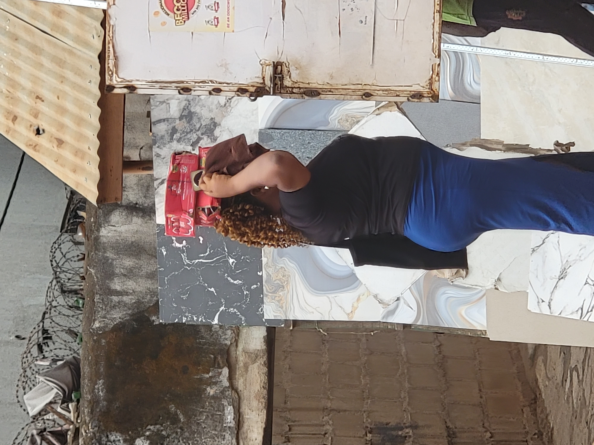 A woman is shown in what appears to be a vendor's stall, possibly in Maiburiji, Nigeria, based on the location provided. She is standing on a display of decorative tiles. The woman is wearing a brown head covering and a black shirt and blue pants. She is holding a package of what looks like red candles. The vendor's stand appears to have a corrugated metal awning overhead with a sign on the side. Barbed wire can be seen near a weathered pole on the left edge of the image.