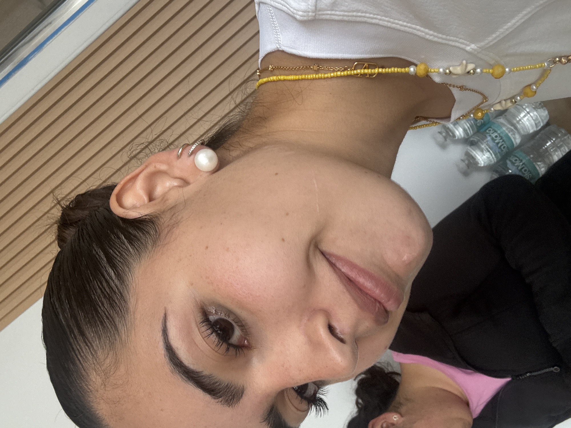 A close-up, angled view of a woman's face. She has dark hair pulled back, light skin, and dark eyes. A pearl earring is visible in her left ear, along with a small silver hoop higher on the ear. She is wearing a white top and a yellow beaded necklace. Partially visible are plastic water bottles. Another person, wearing a pink top and a black jacket, is partly visible on the lower right. The background shows a wooden-slatted wall. Location is not indicated.