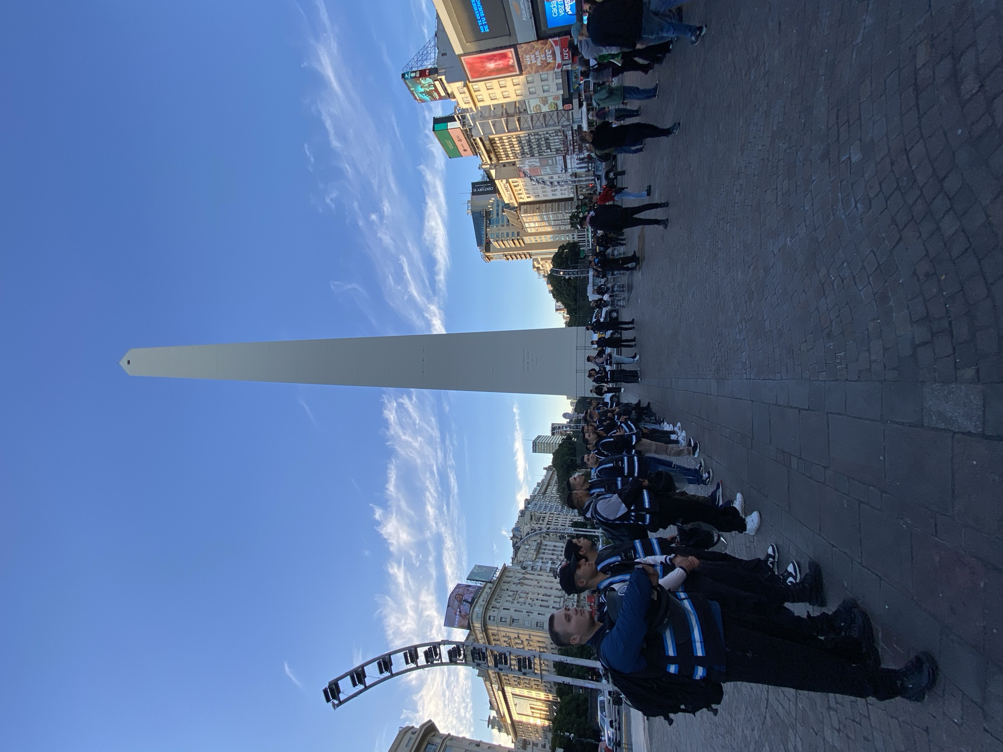 This media file captures a vibrant urban scene in Buenos Aires, Argentina, centered around the iconic Obelisco de Buenos Aires. The tall, white obelisk stands prominently against a clear blue sky, which features scattered, wispy white clouds. The photograph is taken from a street level, looking up towards the monument and the surrounding cityscape.
In the immediate foreground, a group of people is visible, many of whom are wearing distinctive dark blue jackets or vests with light blue and white stripes, suggesting a uniform or coordinated attire. They appear to be standing in a line or formation on a cobblestone surface, attentively facing away from the camera and towards the monument. Other individuals are seen further back, moving about the plaza.
The background is filled with numerous multi-story buildings that line the wide street or plaza. These buildings feature a variety of architectural styles and are heavily adorned with large commercial billboards and digital screens. Visible text on these advertisements includes "JUAN DE GARAY," "CENTURY 21," "KFC," "Atacama," and partial phrases such as "NO TE OLVIDE DE TU SUENG" (likely "Don't forget your dream") and "cada vez n pers" (possibly "increasingly more people").
The lighting suggests either late afternoon or early morning, characterized by a soft, directional light that highlights the monument and buildings, indicating fair weather. The cobblestone ground beneath the people adds to the urban texture of the scene. The overall image portrays a busy public space, typical of a major city, with a group engaged in what appears to be an organized gathering or event.