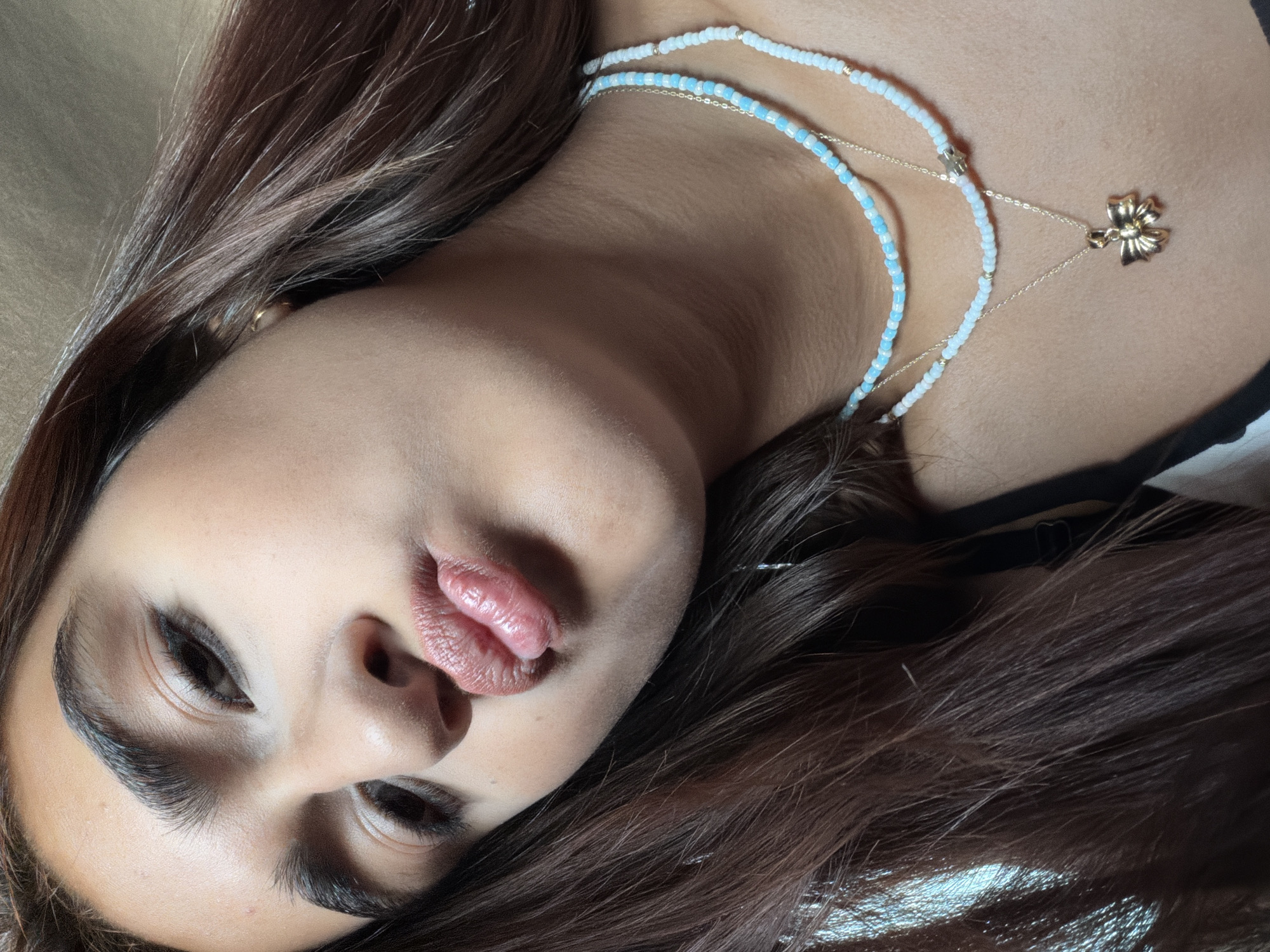 This is a close-up, medium shot of a young woman's face. She is looking down and to the left, with her lips pursed. She has dark, long hair, which is parted to her left and falls around her face. Her eyebrows are dark and well-defined. She is wearing two beaded necklaces, one with light blue and white beads, and another with white and gold beads. The lower necklace has a gold pendant shaped like a bow. The background is a warm, neutral tone, possibly a wall or fabric. There are no other people or objects visible in the frame. The image is brightly lit, with some shadows defining the contours of her face. There is no indication of the city of Shabran, Azerbaijan, in the image.