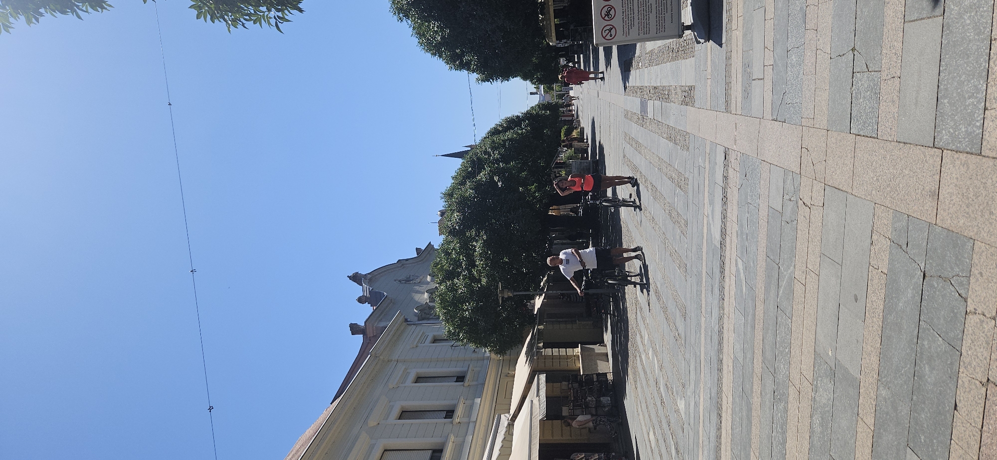 The image depicts a sunny day on a pedestrian street in Keszthely, Hungary. The street is paved with large rectangular grey stones interspersed with narrower pinkish-grey stone strips forming a pattern. Lined with trees and buildings, the scene suggests a commercial or recreational area.
In the foreground, a man in a white t-shirt and dark shorts stands with a bicycle. To his right, a woman in an orange tank top and dark shorts also holds a bicycle. Both appear to be stationary. Further down the street, other pedestrians can be seen walking. On the left side of the street, a building with awnings extends along the sidewalk, with some shops or cafes visible. On the right, a sign with symbols and text is displayed, possibly indicating regulations for the area. The sky is clear blue. The overall impression is of a pleasant, active urban environment.