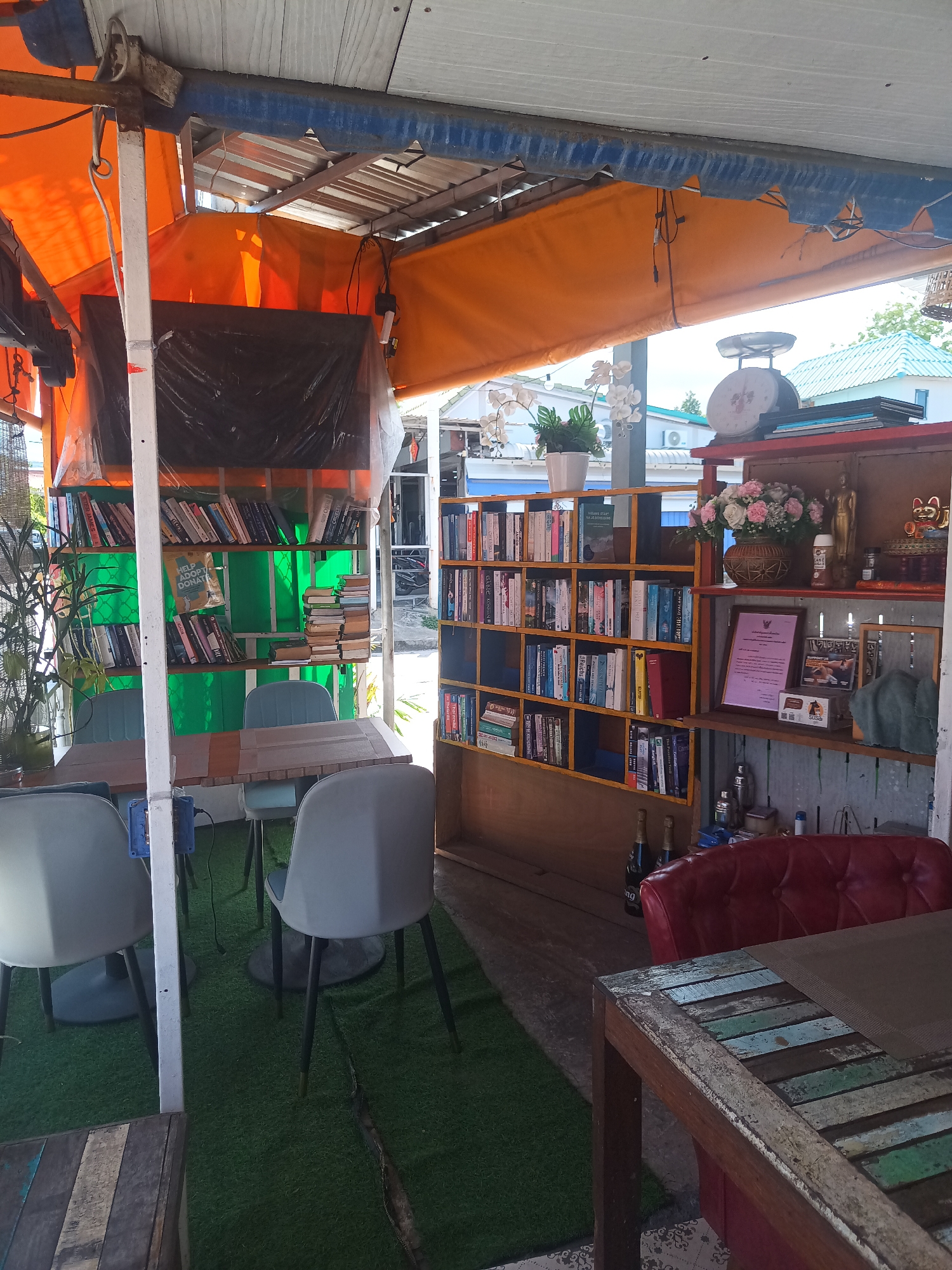 The image depicts the interior of a bookshop or café located in ตำบลเกาะพะงัน, Thailand. Two tables are visible, each surrounded by modern chairs. Bookshelves line the walls, filled with books. A red leather couch is positioned near one of the tables. The floor is covered in artificial green turf. An orange awning covers the area.
