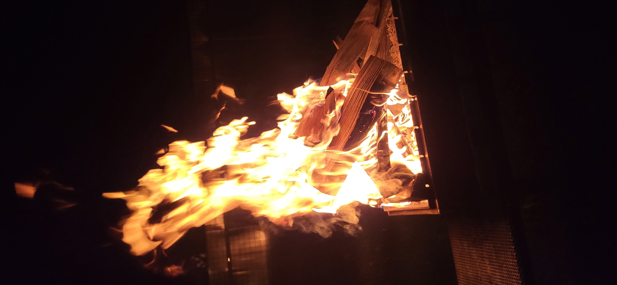 A pyramid-shaped structure of wood is ablaze, emitting intense yellow and orange flames in the dark. The fire is burning vigorously, with flames extending beyond the structure. A faint trail of smoke is visible in front of the fire. The location and city are unknown.