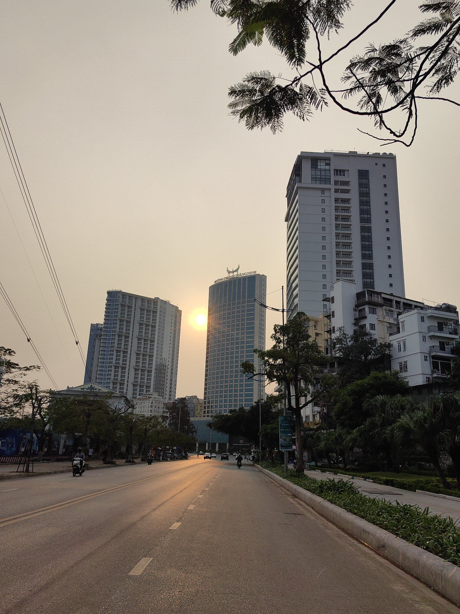 The image depicts a street in Bai Chay Ward, Vietnam at sunset. The street is asphalt and features double yellow lines with a concrete curb on the right. Tall buildings, including the Muong Thanh Quang Ninh Hotel, line the street. Trees are on either side of the street, with some branches visible at the top of the frame. Power lines are visible on the left side of the image. There are some vehicles in the road, including motorcycles. The lighting suggests the sun is setting between the buildings.