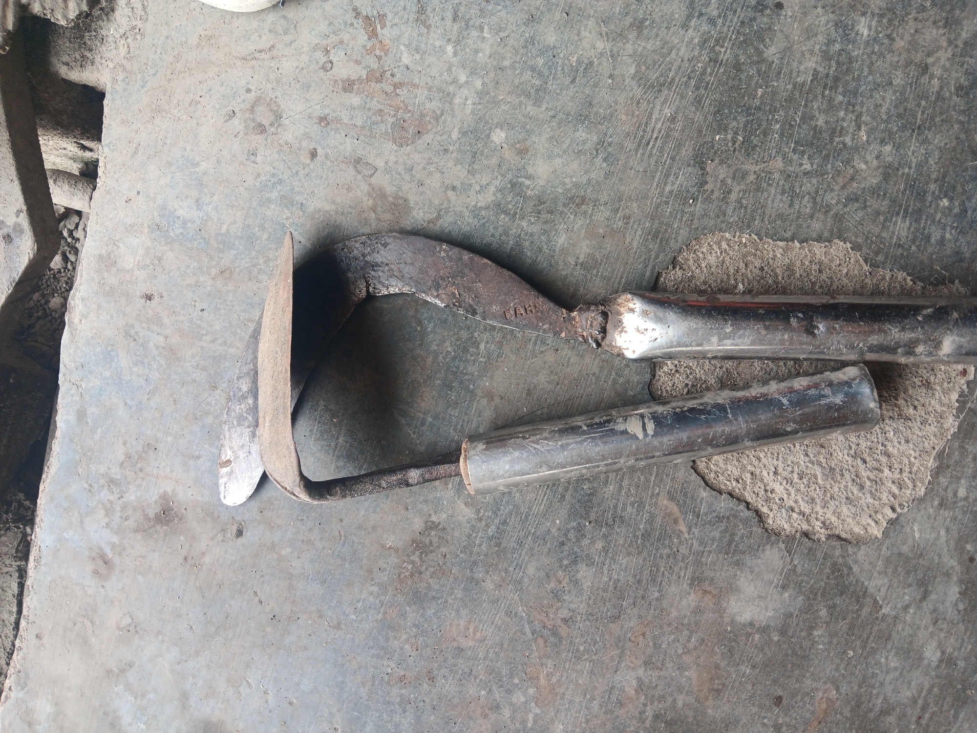 Two gardening tools are laid out on a weathered, grey concrete surface in Batursari, Indonesia. The tool on the left appears to be a hoe, with a broad, curved blade attached to a dark metal shaft that is inserted into a shiny, cylindrical metal handle. The tool on the right is a sickle, characterized by its crescent-shaped, rusty blade with some visible markings that are difficult to decipher. This blade is also attached to a dark metal shaft and inserted into a similar shiny, cylindrical metal handle. The concrete surface shows signs of wear and tear, with cracks and patches of exposed rough material. The overall scene is simple, focused on the tools resting on the ground.