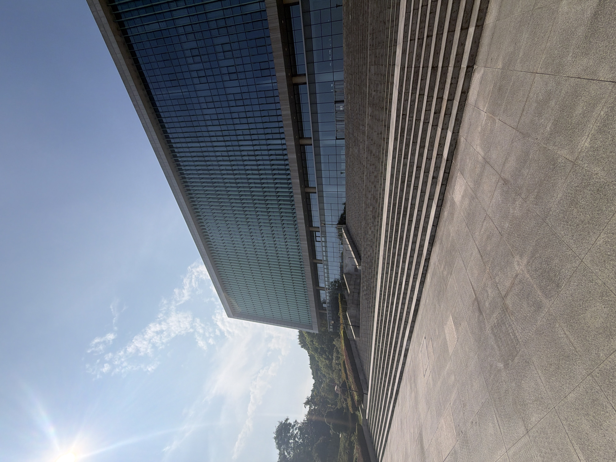 The image shows a wide, low-angle shot of a large, modern building with a glass facade, set against a bright blue sky with scattered clouds. The sun is shining brightly in the upper left corner, casting a lens flare. In front of the building, a wide, grand staircase made of stone leads up to the entrance. The steps are wide and shallow, with a dark stone border. The area at the bottom of the stairs is a paved plaza, also made of stone tiles. The building itself has a long, horizontal design with multiple floors of glass windows. The lower level appears to have a glass entryway with doors. Green trees and landscaping are visible behind the staircase and to the left of the building, suggesting an outdoor setting, possibly a park or campus. The scene appears to be a sunny day. This building is located in the city of Jiangjin, China.