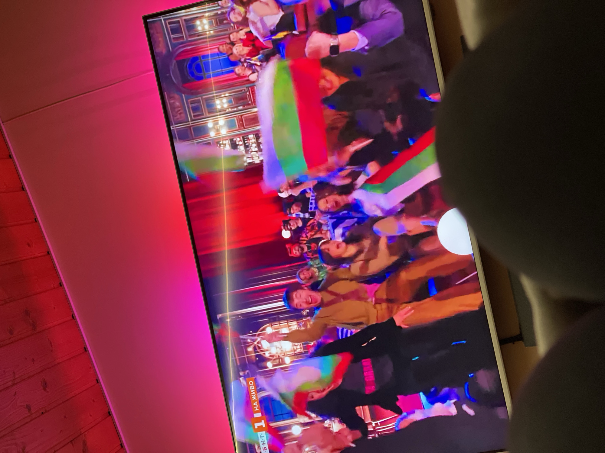 The image shows a television screen displaying a crowded indoor event, likely a celebration. Individuals are visible holding Bulgarian flags. The event appears to be festive. The TV is mounted on a wall with a pink hue emanating from the side of the set. Part of a person's body is blocking the view to the right of the screen. There is a wooden wall on the left. The location in the city of Sofia is not explicitly mentioned within the image, and it is not possible to confidently determine the location based solely on the visual evidence.