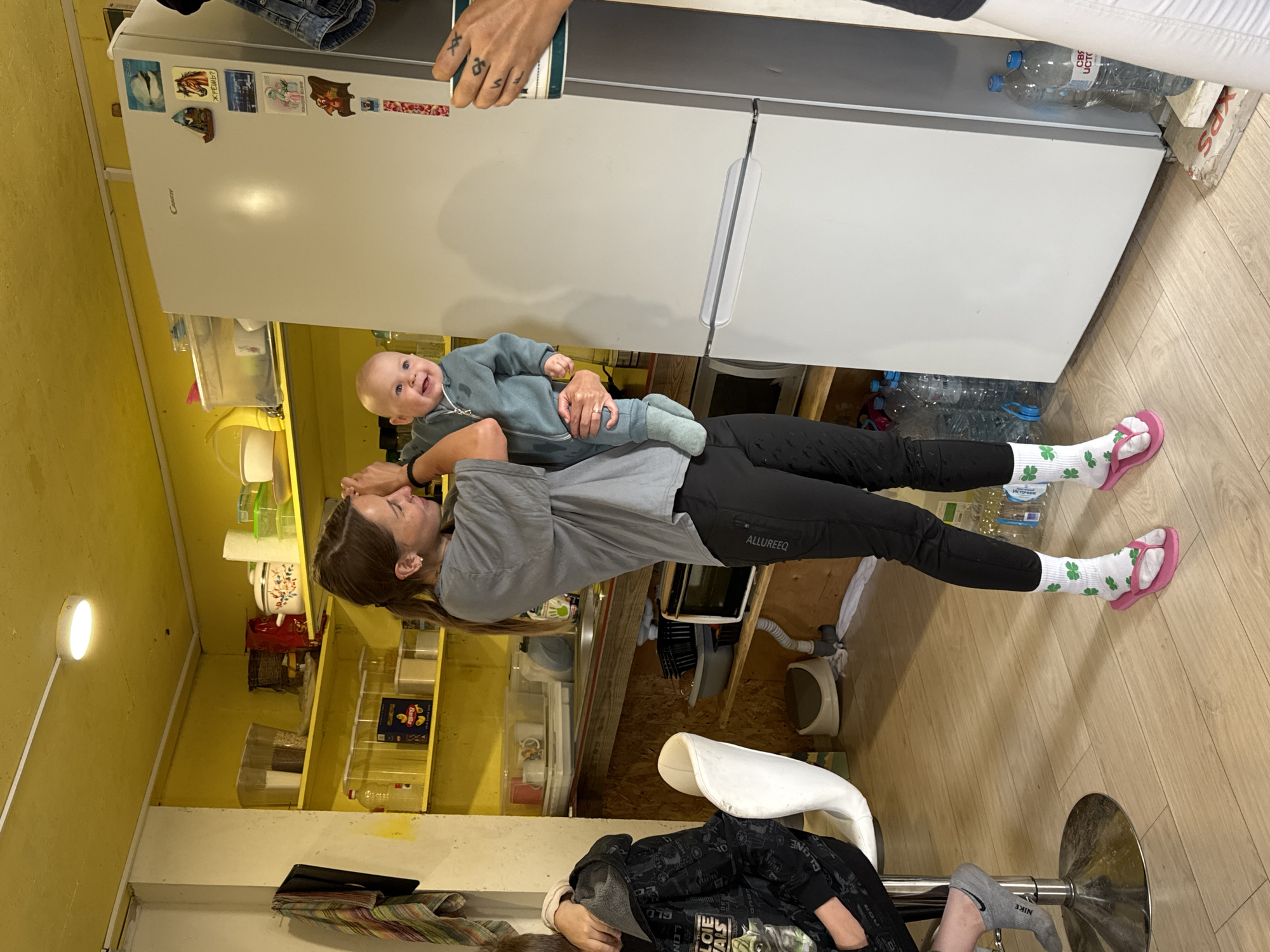 In Dulepovo, Russia, a woman in a grey t-shirt and black leggings is holding a smiling baby in a blue onesie. The woman is standing in a kitchen with yellow walls and shelves filled with various items including food containers, pots, and bottles. A white refrigerator stands to the right of the woman, adorned with several magnets. To the left of the woman, a person wearing a black hoodie is seated on a white stool. The floor is made of light-colored wood. The overall scene depicts a casual indoor gathering.