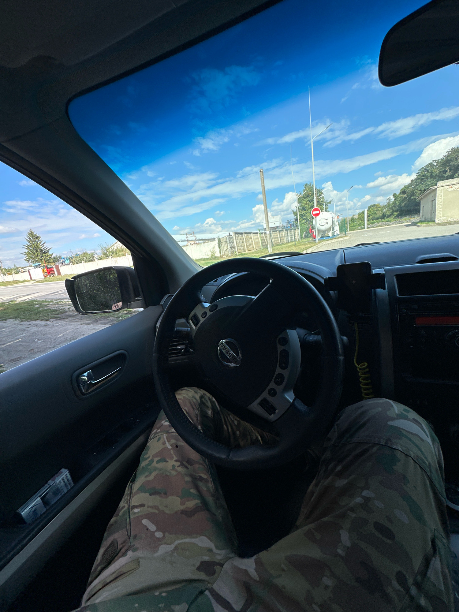 The image shows a first-person perspective from inside a car, looking forward through the windshield. The driver's legs, clad in camouflage pants, are visible in the lower portion of the frame, resting near the steering wheel. The steering wheel is a black Nissan model. Outside the car, the sky is blue with scattered white clouds. In the distance, there is a road, a few trees, some buildings, and what appears to be an industrial area with structures like gas tanks and a red tractor. There are also power lines and streetlights visible. The specific location is identified as Shatava, Ukraine. The interior of the car includes a side door with a handle and a car charger or phone mount. A pack of cigarettes is visible in the door pocket. The overall scene depicts a person sitting in a parked vehicle on a clear day, with a view of both the car's interior and the surrounding environment of Shatava, Ukraine.