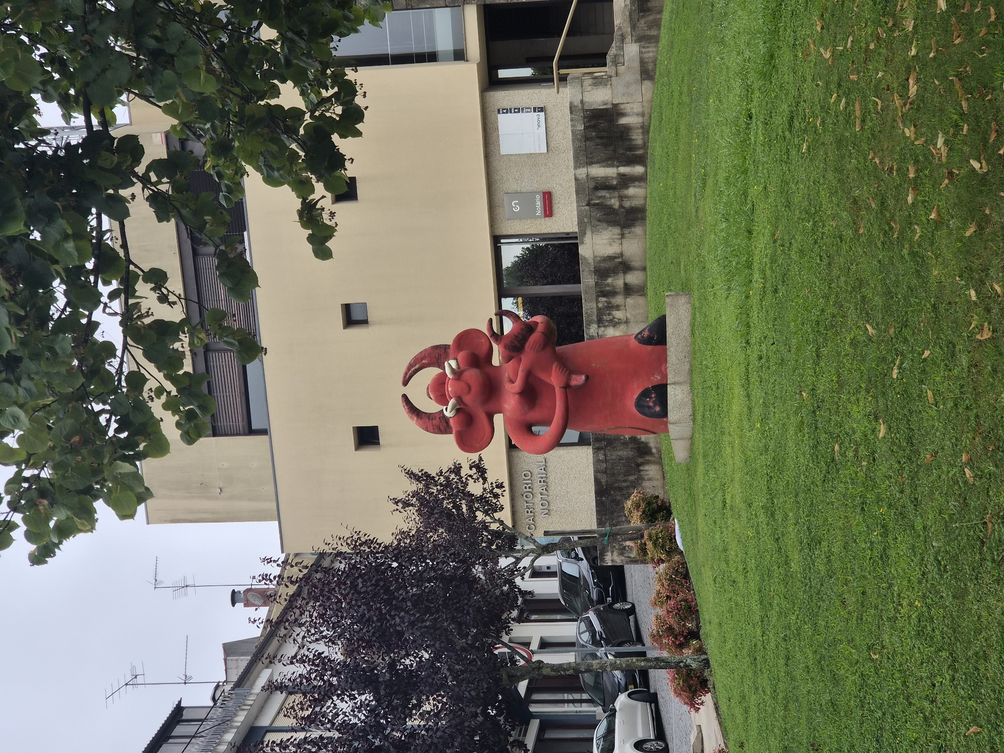 The image captures an outdoor scene in front of a modern building with a beige facade. In the foreground, a large, vibrant red abstract sculpture resembling a stylized bull holding a smaller figure is prominently displayed on a grassy area. The sculpture has large horns, wide eyes, and a whimsical, somewhat childlike representation. Behind the sculpture, the building features large windows and a sign that reads "CARTÓRIO NOTARIAL". To the right of this sign, other signs are visible, one with the word "Notario" and a logo, and another white sign with text. The area is landscaped with green grass, some bushes with red and pink foliage, and trees with dark purple leaves on the left side of the frame. In the background, several cars are parked on a cobblestone street alongside the building. The sky is overcast, suggesting a cloudy day. This scene is located in the city of Barcelos, Portugal, specifically in front of a notary office.