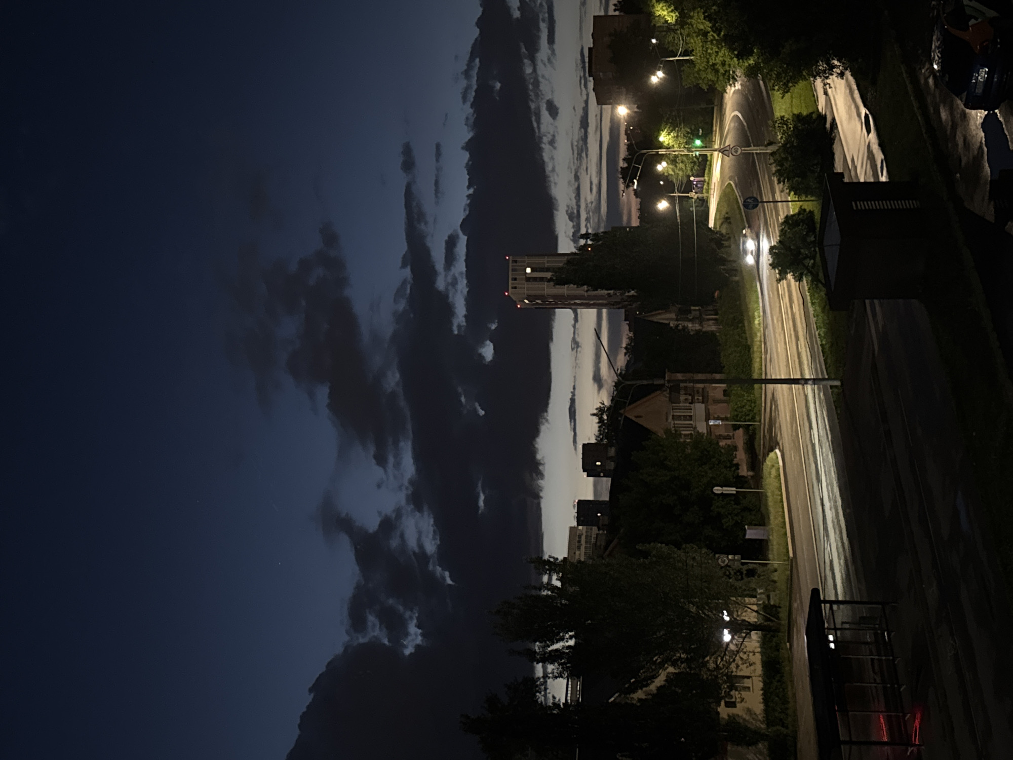 The image presents an urban street scene at night or dusk, viewed from an elevated perspective. The sky, dominating the left half, is a dark blue-grey, filled with dramatic, elongated clouds. A lighter strip of sky is visible towards the horizon on the upper right.
The central subject is a wet, winding road, glistening with reflections from numerous streetlights that illuminate its surface and the surrounding trees. White road markings are visible on the asphalt. A single vehicle with bright headlights is seen driving on the road, heading away from the viewer.
Along the road, several streetlights are on, casting warm yellow and white light. Lush green trees line the street and are interspersed among buildings, some brightly lit, others appearing as dark silhouettes. In the mid-ground, a prominent tall, light-colored building with multiple windows and red lights on its rooftop structure stands out. Other smaller buildings are visible further in the background.
Visible street furniture includes a tall, slender pole-like structure near the tall building. Road signs are also present: a triangular sign with a red border clearly shows the number "40" (indicating a speed limit), and a circular blue sign is visible below it. The foreground includes a dark, rectangular structure, possibly part of a balcony or railing.
The wet road suggests recent rain or damp conditions. The scene is quiet and still, capturing a calm moment in an urban environment at the transition from day to night. No people are explicitly visible. The setting is consistent with an urban area like Perm, Russia, as indicated by the context.