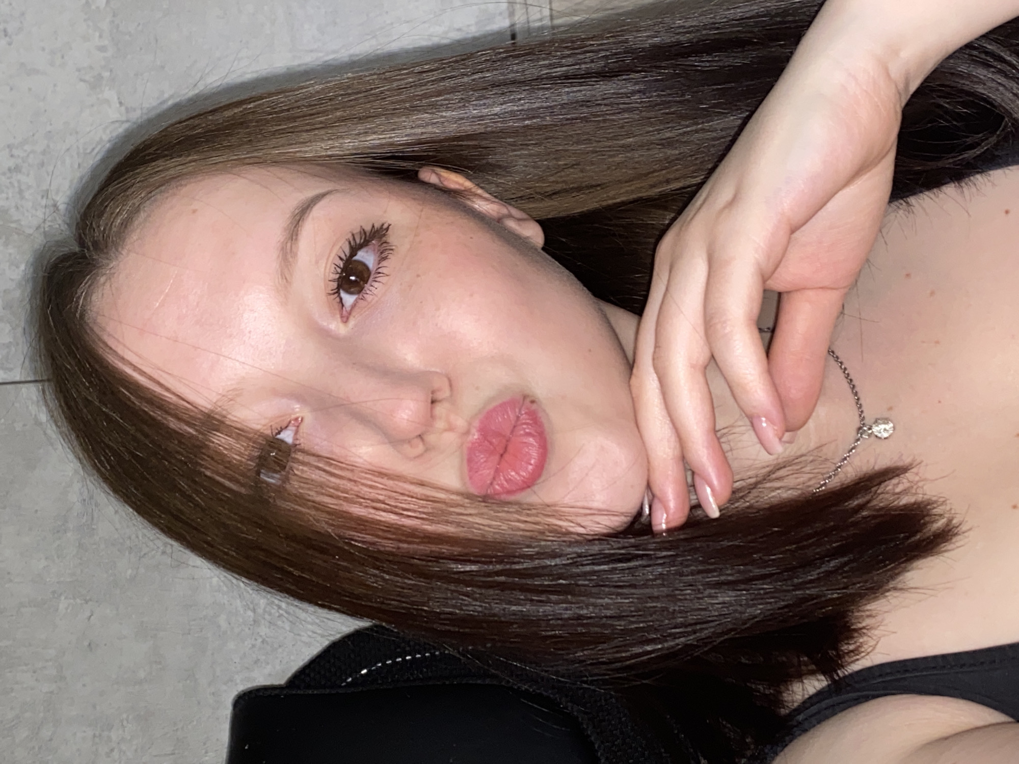 A close-up photograph depicts a young woman with light skin, brown eyes, and brown hair. She is reclining, with her face tilted towards the camera and her lips pursed. Her right hand is positioned near her neck. She is wearing a black top and a delicate silver necklace. The background appears to be a light gray wall. No information suggests the location of Chaikovsky, Russia.
