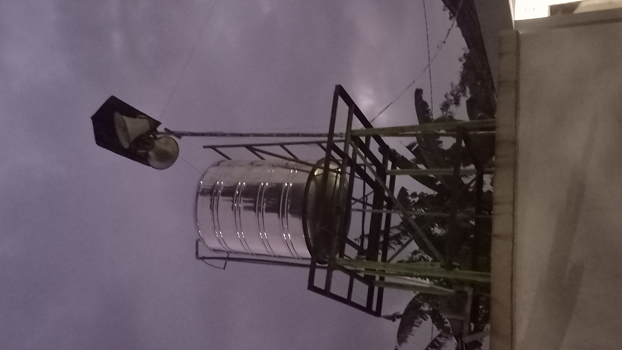 In the city of Pare, Indonesia, a cylindrical stainless steel water tank stands on a metal platform. Above the tank, a street lamp with a black shade is mounted on a pole. The scene is set against a cloudy, twilight sky. The water tank is supported by a green metal structure, and its shiny surface reflects the ambient light. Banana trees are visible behind the structure. The foreground consists of a light-colored concrete wall, with shadows cast upon it.