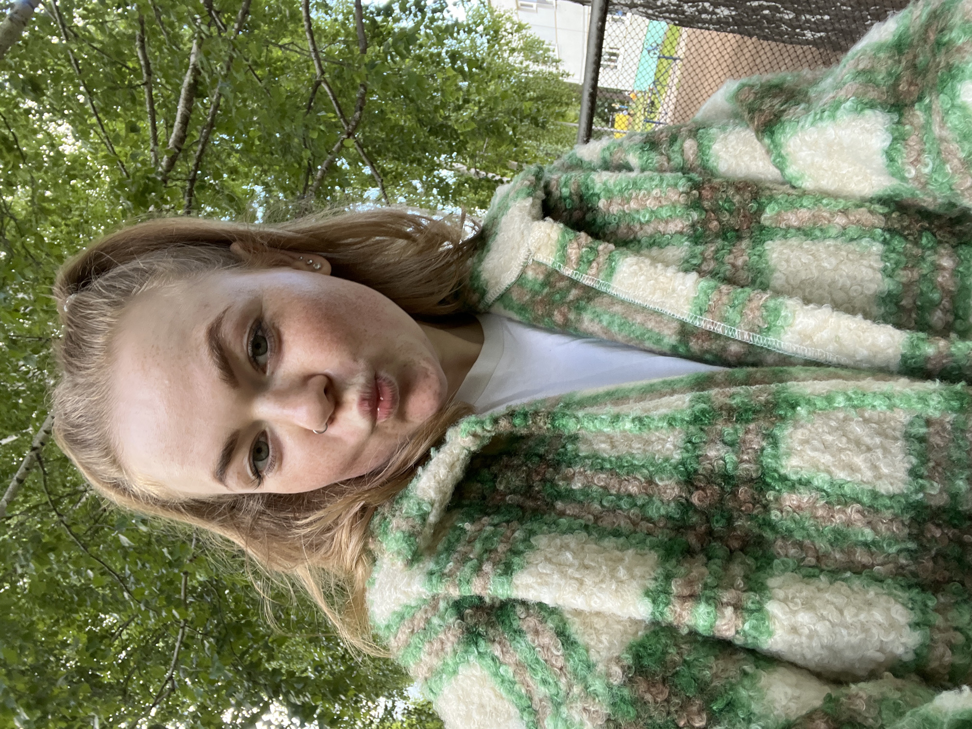 A young woman with light blonde hair, fair skin, and light-colored eyes is captured in a close-up, low-angle portrait, looking directly at the camera with a playful, slightly puckered expression. She has a small silver nose ring on her left nostril. She is wearing a white undershirt and a prominent, textured jacket or coat. The jacket features a distinct, irregular plaid pattern in shades of light green, darker green, cream/off-white, and light brown, made of a fluffy, bouclé-like material.
The scene is set outdoors in Chaikovsky, Russia, during daylight hours, indicated by the bright, natural light filtering through abundant green foliage. The perspective is from below, looking upwards into the canopy of deciduous trees, whose leaves are in full bloom, suggesting a spring or summer setting. In the upper right background, beyond the trees, there is a blurry hint of an urban structure, possibly a fence or building, and a light-colored sky. The lighting is bright and even, highlighting the details of the woman's face and the texture of her jacket. There is no visible text in the image.
