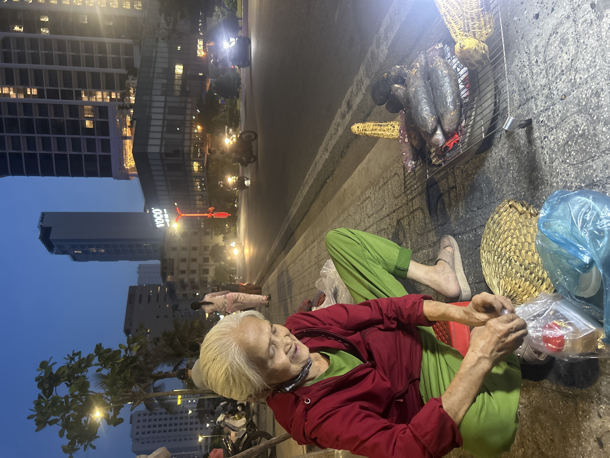 The image captures a street scene in Bắc Nha Trang, Vietnam at night. An elderly woman sits on a red plastic stool on a concrete sidewalk. She wears green pants, a green shirt, and a red jacket. She is occupied with packing food items in a clear plastic bag, with a woven basket and other plastic bags next to her.
Beside her is a small charcoal grill on the sidewalk, containing roasted corn and sweet potatoes. A building with the brand "VOCO" is visible in the background. Streetlights illuminate the scene. Motorbikes are parked nearby and in the background, and other pedestrians walk along the sidewalk. Motor vehicles move along the road.