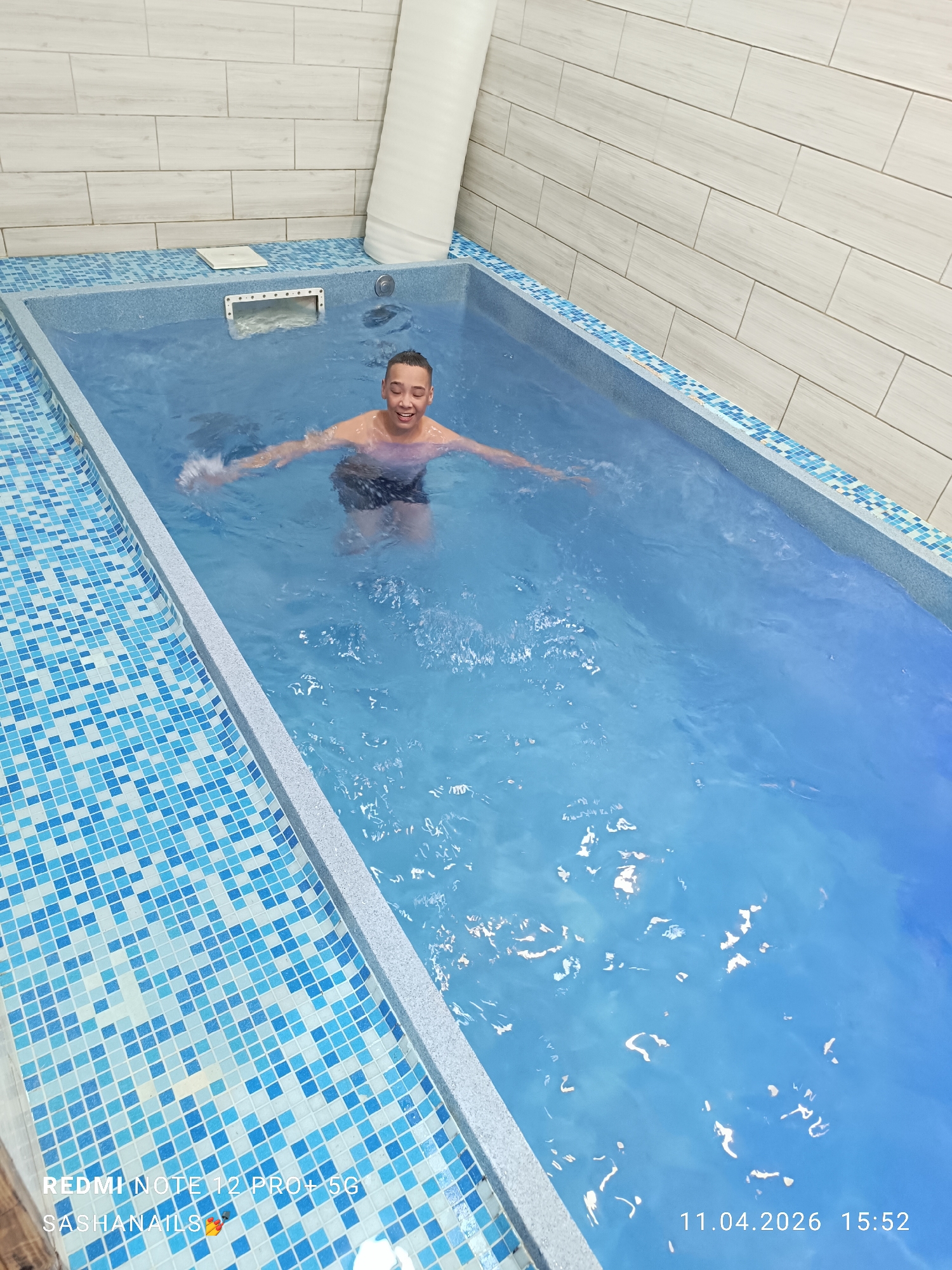 A young person is in a rectangular, indoor swimming pool. The person is standing, with arms outstretched to either side, a smile on their face. The pool is lined with blue and white mosaic tiles and has a gray border. The walls of the room are tiled in light gray, brick-like tiles. A white, cylindrical object is visible in the corner of the room behind the pool. The timestamp "11.04.2026 15:52" is present in the bottom right corner of the image. The image origin is identified as "REDMI NOTE 12 PRO+5G" with "SASHANAILS" also present. There is no clear indication of the city Chaikovsky, Russia in the image.