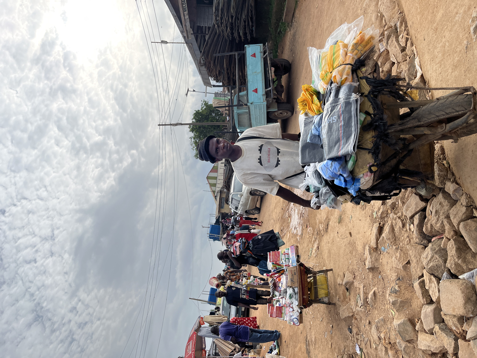 This media file captures a street scene in Gura Top, Nigeria, likely during the day with overcast skies. The primary subject is a young man standing next to a cart piled high with goods, predominantly textiles and items wrapped in plastic bags. He is wearing a white t-shirt with a graphic design and a dark beanie. His gaze is directed towards the camera with a neutral expression.
The cart itself is a makeshift affair, appearing to be a modified bicycle or motorcycle with a platform attached, laden with yellow and gray fabric bags, some with visible text like "SHS". The goods are secured with black plastic ties. Behind the man and his cart, the scene opens up to a bustling market or roadside with various other vendors and stalls. A light blue truck carrying what looks like construction materials is parked nearby, and several other vehicles are visible further down the road.
People are milling about, suggesting ongoing commercial activity. There are glimpses of other stalls displaying a variety of items, including what appear to be cosmetics, toiletries, and other small goods. The ground is a mix of dirt and scattered rocks, indicative of an outdoor marketplace. The sky is filled with textured clouds, hinting at a potentially humid or pre-rainy weather condition. The overall impression is one of everyday life and commerce in a Nigerian setting.