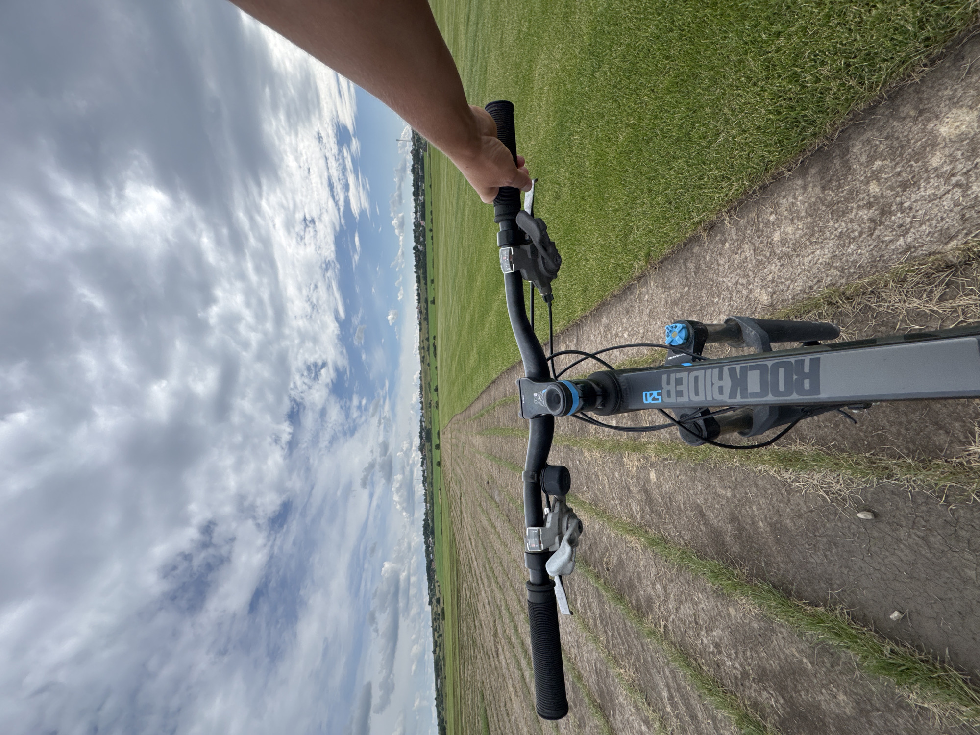 A first-person perspective shot shows a person's arm and hand gripping the handlebars of a bicycle. The bicycle is on a dirt path that runs through a large, flat green field. Rows of young green plants are visible in the field, separated by dry soil. In the distance, a line of trees and buildings can be seen, suggesting a settlement. The sky is cloudy with patches of blue. The image appears to be taken in Kempelevo, Russia, on a daytime.