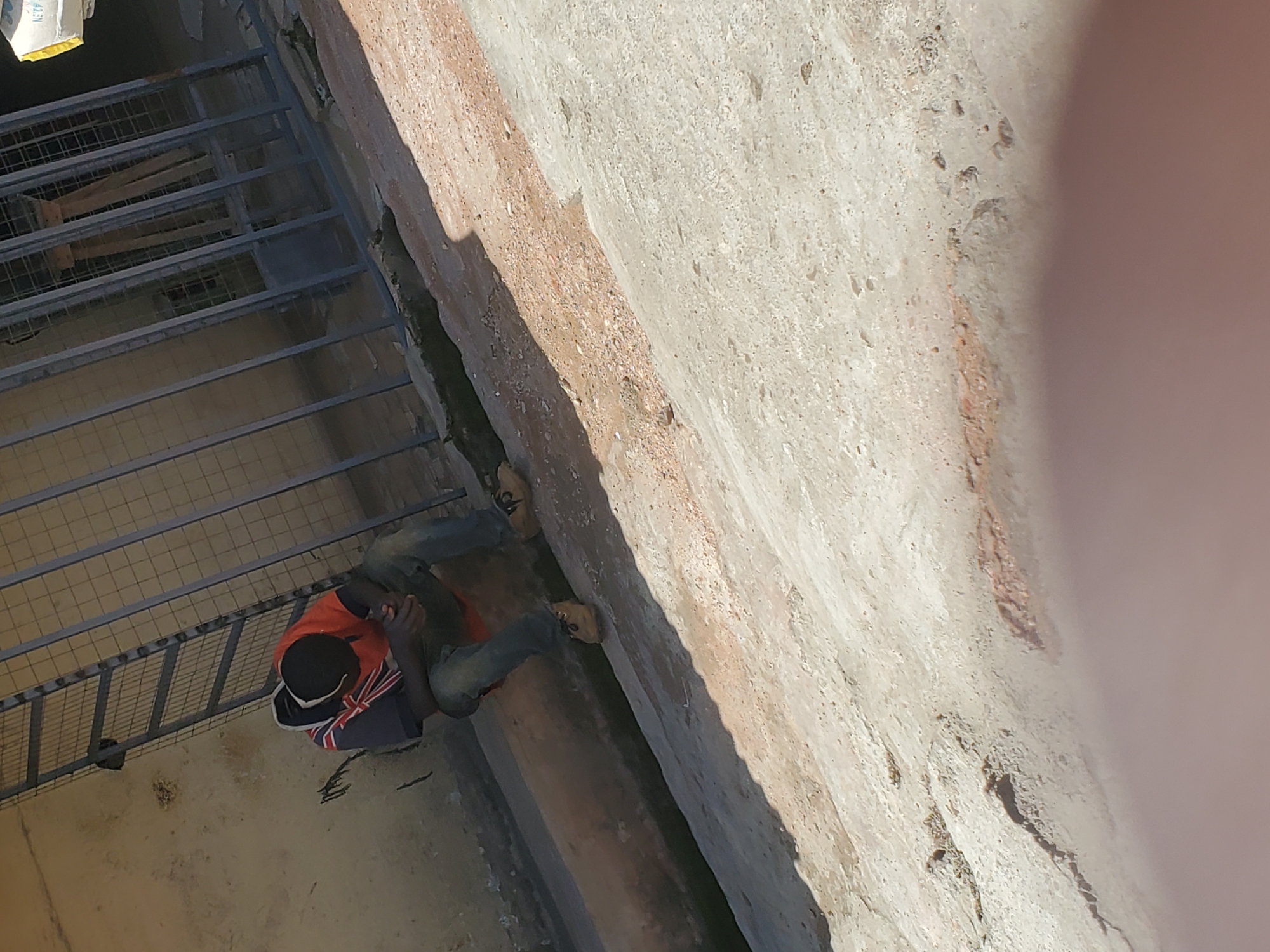 A Black male, appearing to be in his late teens or early twenties, is sitting on a concrete ledge, facing away from the camera, with his head bowed. He is wearing a dark hooded shirt with a British flag design on the front, an orange t-shirt underneath, blue jeans, and tan shoes. His hands are clasped in his lap. He appears to be alone. The background consists of a textured, yellow wall and a metal fence. The ground in front of him is a mix of dirt and gravel, with a concrete channel running along the wall. The lighting suggests it is daytime, with shadows indicating the sun is high. The scene is outdoors in an urban environment. The presence of the British flag design on the clothing may suggest a connection to the UK or its cultural influence. The specific location of Bukuru, Nigeria, indicates a West African setting.