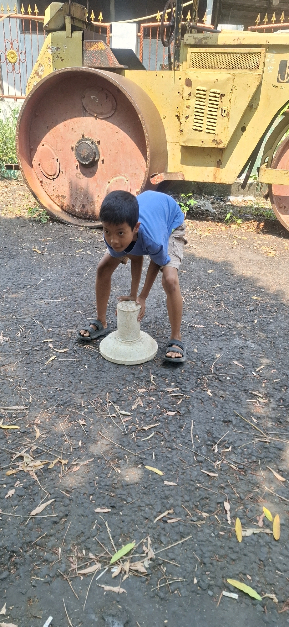 A young boy wearing a blue t-shirt and khaki shorts is crouched down on an asphalt surface, holding onto a light-colored, cylindrical object with a flared base. He is positioned in front of a large, yellow, and rusty piece of construction equipment, possibly a road roller, with a prominent reddish-brown drum. The background shows a partial view of a metal fence with yellow decorative elements and some green foliage. The ground is covered with scattered dry leaves and twigs. The scene appears to be outdoors, possibly in a yard or open area within the city of Cilaku, Indonesia, given the context of the image. The boy's body is angled, with his knees bent and his head looking forward, suggesting he is engaged with the object he is holding. The lighting indicates it is daytime.