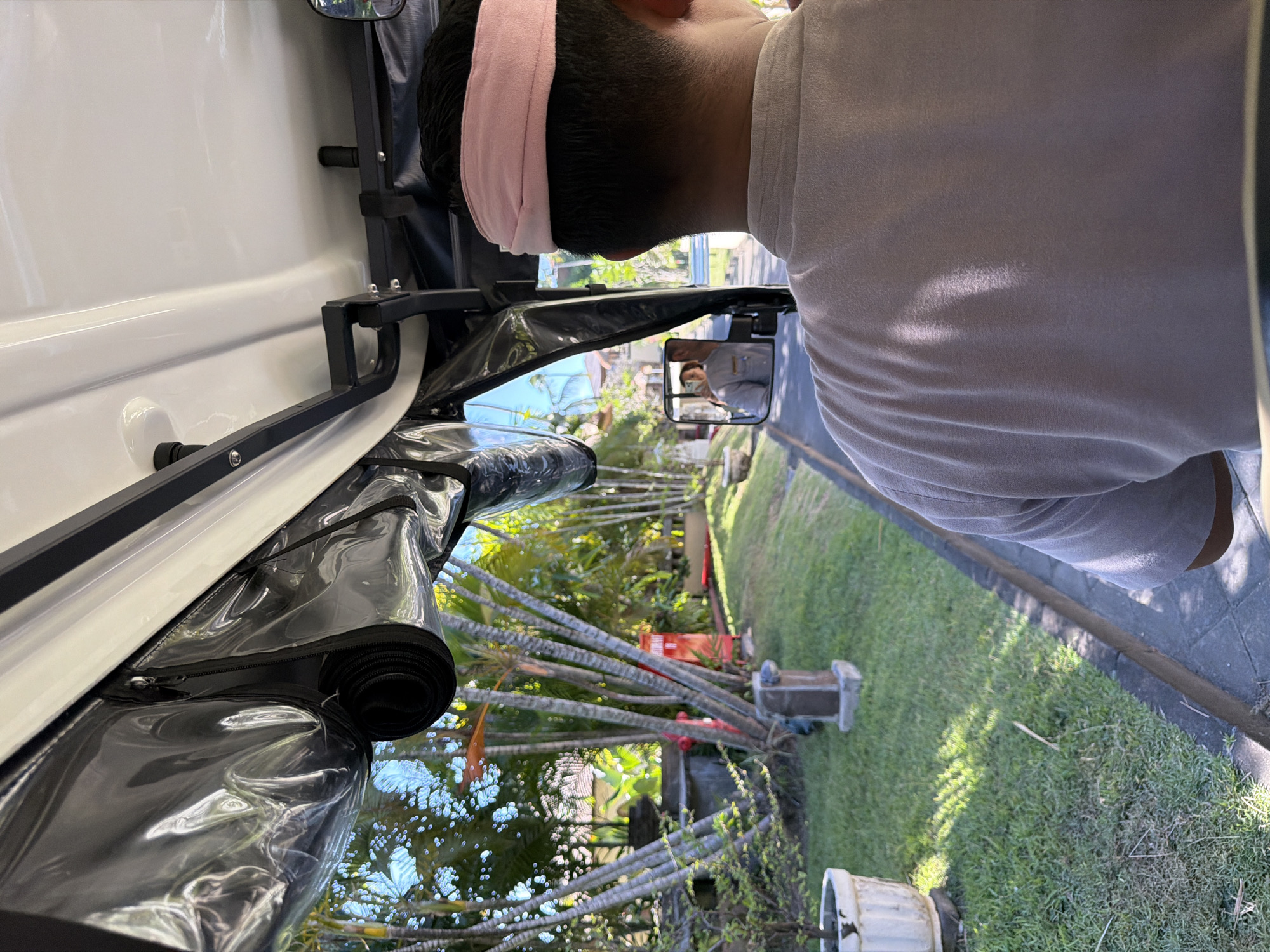 An outdoor scene captured in bright daylight shows a person from behind, seated in what appears to be an open-sided electric vehicle or golf cart. The person wears a light-colored short-sleeved t-shirt and a pink headband, with short dark hair visible beneath.
The vehicle features white body panels and a black frame structure, possibly a roll bar or canopy support. Clear plastic coverings, designed to enclose the side of the vehicle, are seen rolled up along the top edge, secured with black straps and zippers. A side mirror on the right reflects another person, likely the same individual or a passenger, wearing a light top and possibly holding a phone or camera.
The setting is a lush, tropical environment, consistent with Kuta, Indonesia. A well-maintained green lawn spans the right side of the image, bordered by a paved path or curb. The background is dominated by numerous tall, slender palm trees and dense tropical foliage, suggesting a garden, resort, or residential area. Scattered among the greenery are a few decorative garden elements or plant pots, and a red object is partially visible behind the trees.
The clear, sunny weather is evident from the strong shadows cast on the grass and the vehicle. The scene depicts a moment of observation or transit through a verdant landscape. No visible text is present in the image.