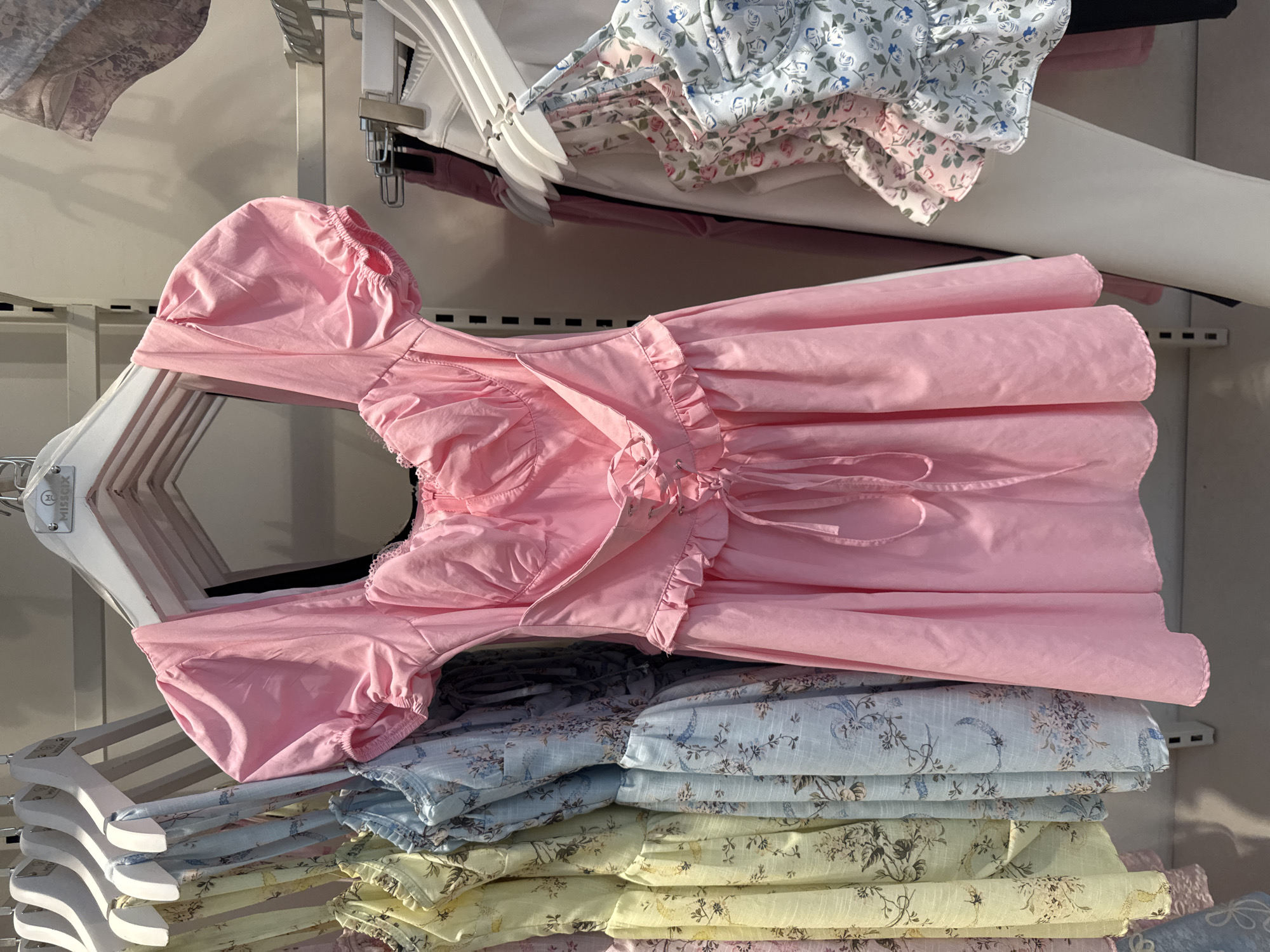 The image shows a clothing store in Gungoren, Turkey. In the foreground, a pink dress with short puff sleeves, a square neckline, and a lace-up corset detail is displayed on a hanger. The dress has ruffled trim at the hem of the skirt and around the corset. To the left of the pink dress, several other dresses in pastel colors with floral prints are hanging. To the right, a white garment and a floral camisole are visible. The dresses are displayed on white hangers against a light-colored background with metal shelving units. The store appears to be well-lit and organized.