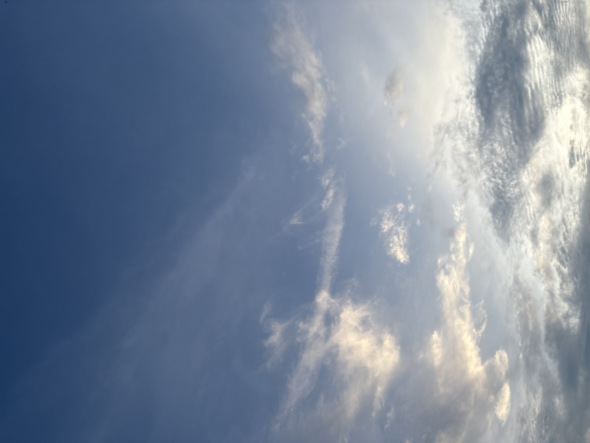 The image depicts a wide-angle view of a sky with scattered clouds. The sky is a gradient of blues, deepening to a darker shade at the top and becoming lighter towards the bottom. wispy, thin clouds are spread across the upper half of the frame, appearing white and translucent. In the lower half, denser clouds are visible, some tinged with yellow and white from sunlight. These clouds exhibit more texture, with some appearing layered and others resembling small, puffy formations. The overall impression is that of a daytime sky, possibly during the late afternoon or early morning, with the sun's light filtering through the clouds. There are no discernible buildings, people, or specific landmarks visible, suggesting an open-air perspective. The image does not provide any direct clues to its location within North Port, United States of America; it is a generic sky scene.