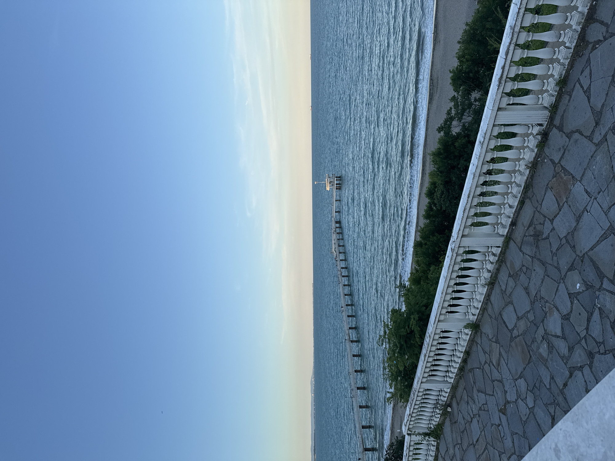 This image depicts the Burgas Sea Garden, specifically the pier extending into the Black Sea. The scene is captured from an elevated vantage point, looking out over the water. In the foreground, a stone-paved walkway is visible, bordered by a white balustrade with ornate pillars. Beyond the balustrade, lush green trees and bushes line the edge of the beach. The sea is a deep blue, with gentle waves lapping the sandy shore. A long pier stretches out into the sea, supported by numerous pillars. People can be seen walking on the pier, and at its end, there is a small, white, enclosed structure with a light post. The sky is clear and blue, with a gradient towards a lighter hue near the horizon, suggesting it is late afternoon or early evening. Several ships are visible in the distance on the horizon.