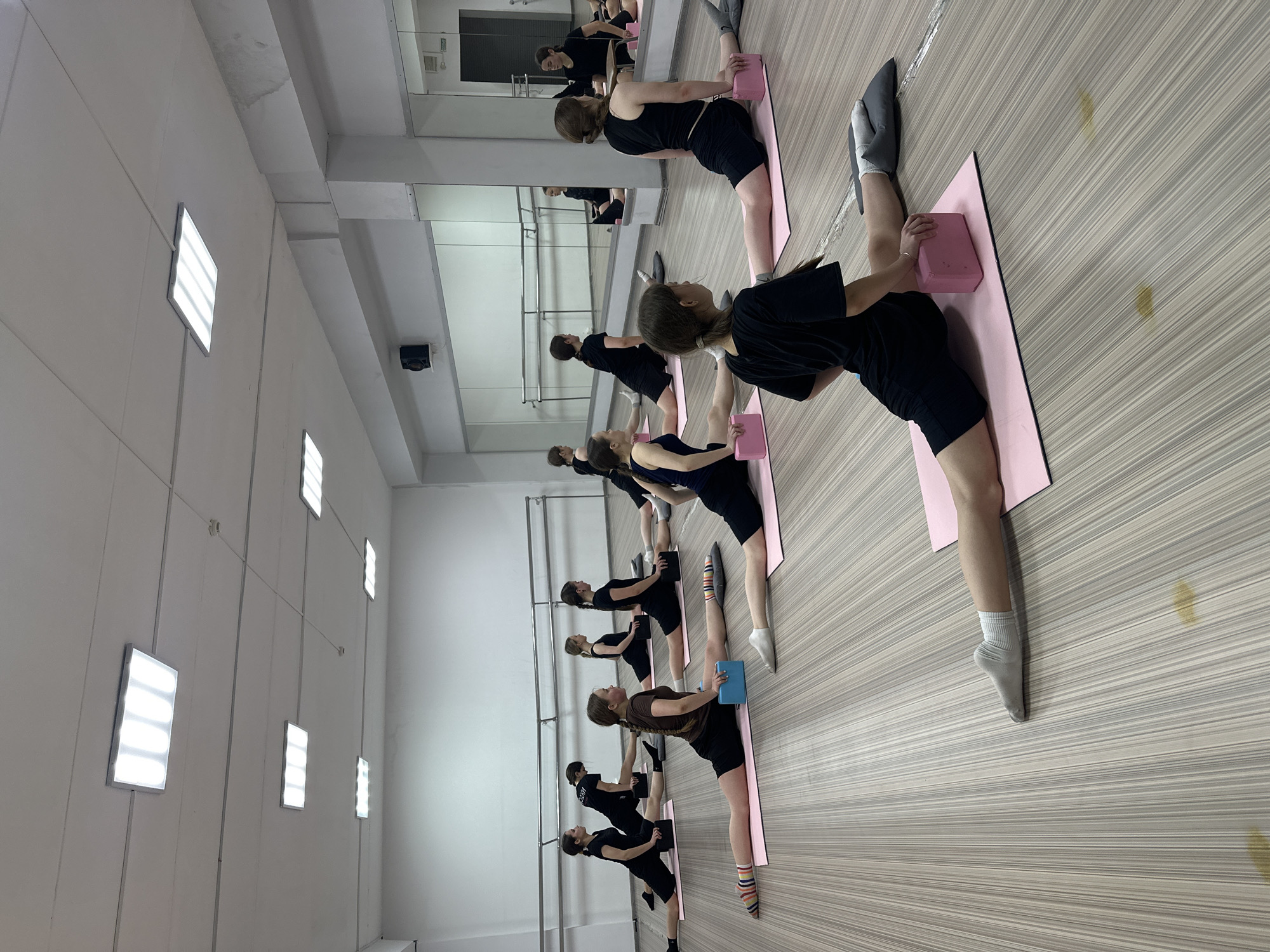In a brightly lit dance studio in Votkinsk, Russia, a group of approximately eight young women are engaged in a stretching exercise, likely ballet-related. They are arranged in a line, each positioned on a pink or blue yoga mat with a yoga block in hand. The women are dressed in dark-colored athletic wear, some wearing socks or ballet shoes. The studio has a light-colored wood floor and a mirrored wall on the right side, which reflects the participants and expands the perceived space. Linear lighting fixtures are arranged along the wall on the left, and the walls are white. The overall scene suggests a training environment, with focus on flexibility and technique.