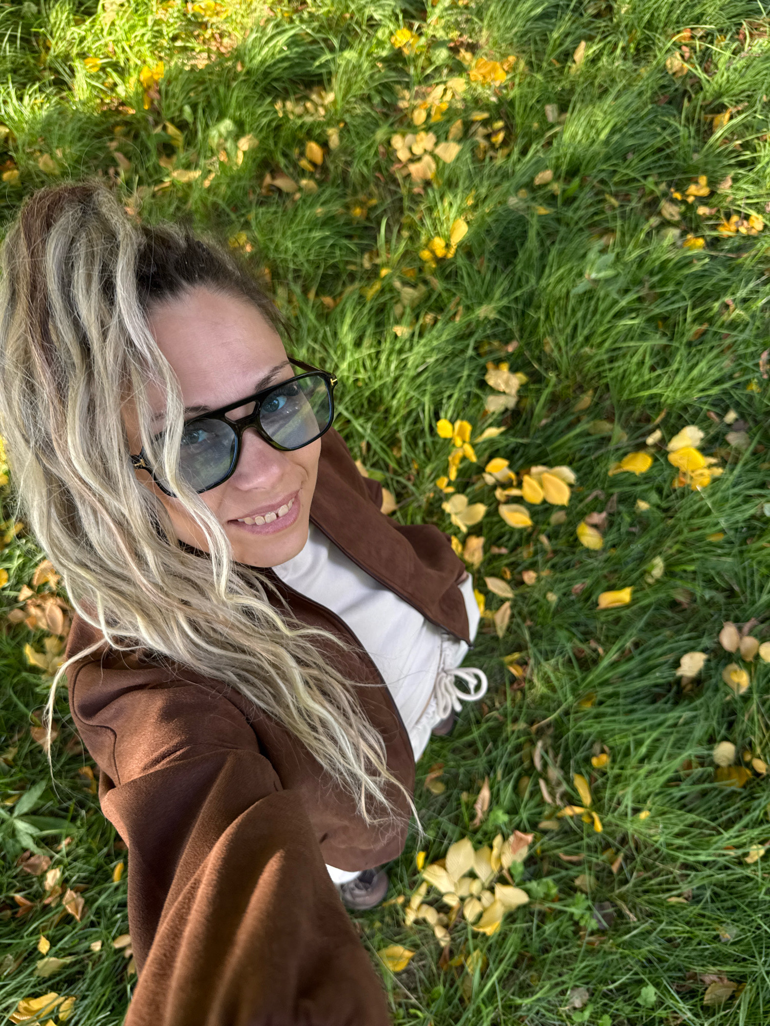 The image shows a selfie taken from above of a woman with blonde, wavy hair tied in a high ponytail. She is wearing large black-framed glasses and a brown jacket over a white garment. She is smiling at the camera. The woman is standing in a grassy area with scattered yellow leaves, indicating an autumn setting. The background consists of green grass, some of which is illuminated by sunlight, creating patches of light and shadow. The location is identified as Rusanovo, Russia.