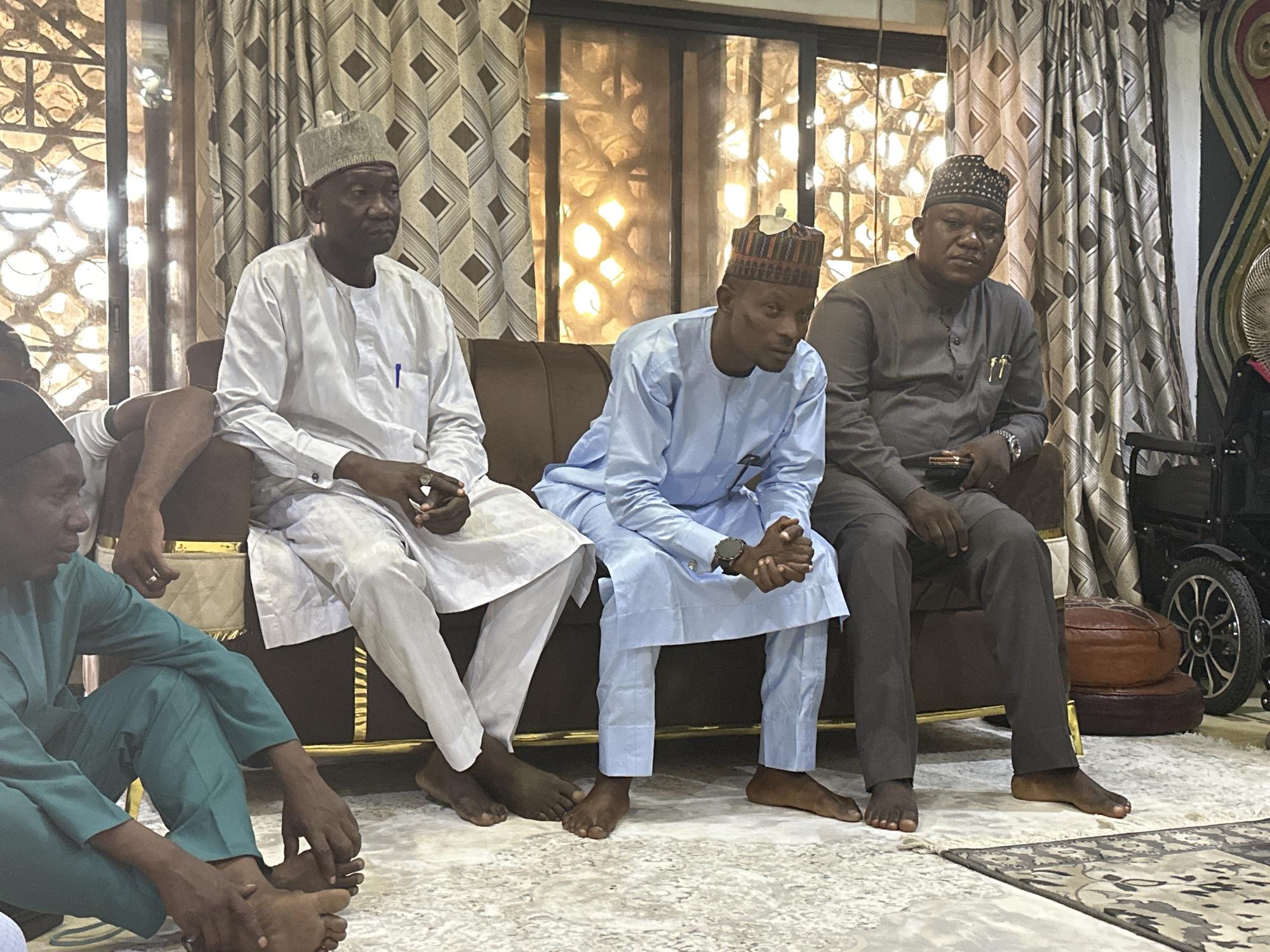 In an indoor setting in Unknown, Unknown, three men are seated on a brown couch. The man on the left wears a white outfit and a grey hat. The man in the middle is dressed in light blue attire with a colorful hat. The man on the right is in a grey outfit and a dark hat. All three men are barefoot. To the left, partially visible, are other people dressed in green outfits. Behind the couch, there is a patterned window and draped curtains. A wheelchair is visible on the right side of the image. The floor is covered with a patterned rug.