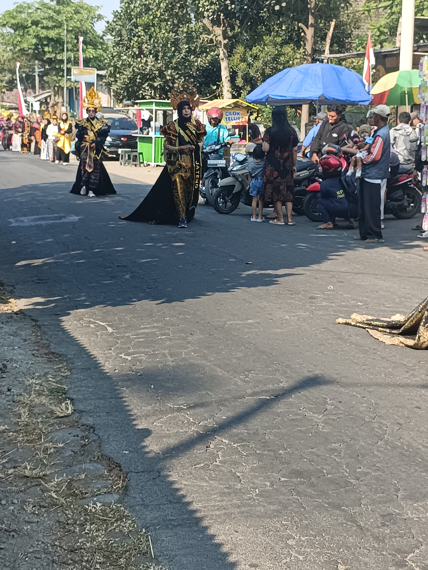 In Jatitengah, Indonesia, a parade is underway on a paved street under a clear sky. Two individuals in elaborate traditional attire, adorned with gold headpieces and ornate dark robes, walk down the center of the road. The person on the left wears a tall, multi-tiered gold headdress and a black robe with gold accents and gold fabric draped over their arms. The person on the right wears a similar gold headdress and a dark robe with intricate gold patterns and a sash that reads "JATENG."
A line of people in colorful clothing, likely participants or onlookers, stretches down the left side of the street, some carrying flags. Behind them, a black car is parked. To the right of the parade, several motorcycles are parked. A blue umbrella provides shade for a group of people gathered around the motorcycles. Among this group, a man in a black hoodie is smoking, and a man in a blue shirt is on a motorcycle. Another person in a dark uniform is crouched down next to a motorcycle. Other individuals, including a child being held by an adult, are visible in this area.
In the background, trees line the street, and a few stalls with awnings are set up. One stall has a sign that reads "CILOK TELUR." The street pavement is cracked and worn in places. Shadows cast by the trees and possibly overhead structures create patterns on the road. The overall scene suggests a festive community event taking place in a village or small town.