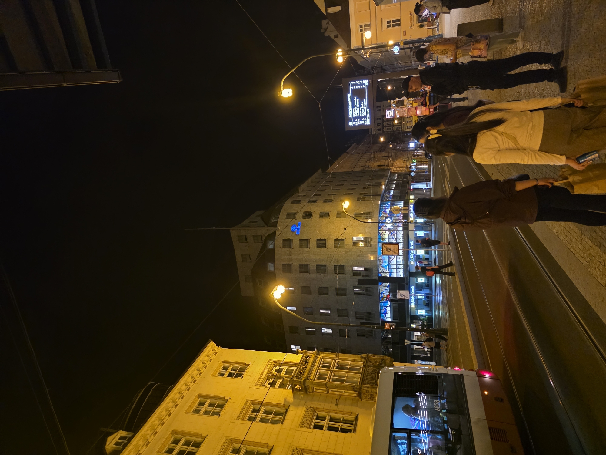 An urban street scene unfolds at night, illuminated by numerous artificial lights. The perspective is slightly elevated, looking down a street or plaza flanked by buildings.
In the foreground, a set of tram tracks runs down the right side of the street. A white and brown tram, marked with the number "6878," is partially visible on the far right. Adjacent to the tram tracks, a sidewalk hosts several pedestrians. Closest to the viewer, a person in a dark brown jacket and black trousers stands with their back mostly turned. Further back, two individuals walk away from the viewer: one wearing a light-colored top and brown pants, carrying a light bag, and another in a darker top with a yellow hair clip. Beyond them, a person in black stands near a digital display board, which shows tram information in Czech, including phrases like "Pravidelnost jízd" (Schedule regularity), "zastávek" (of stops), "na lince" (on line), "Zpoždění" (Delay), and "Služby" (Services), along with a time "00:24."
Buildings line both sides of the street. On the left, a multi-story building with ornate architectural details is bathed in warm yellow light from ground-level streetlights. Its windows glow with internal light. In the mid-ground, a taller, grey building features a prominent blue cross symbol and a "Hilton" sign. More commercial signs, some illuminated, are visible along the street, though their text is mostly unreadable. Overhead, power lines or cables crisscross the dark night sky. The overall impression is a bustling city environment after dark, likely in a Central European location given the architectural style and readable Czech text.