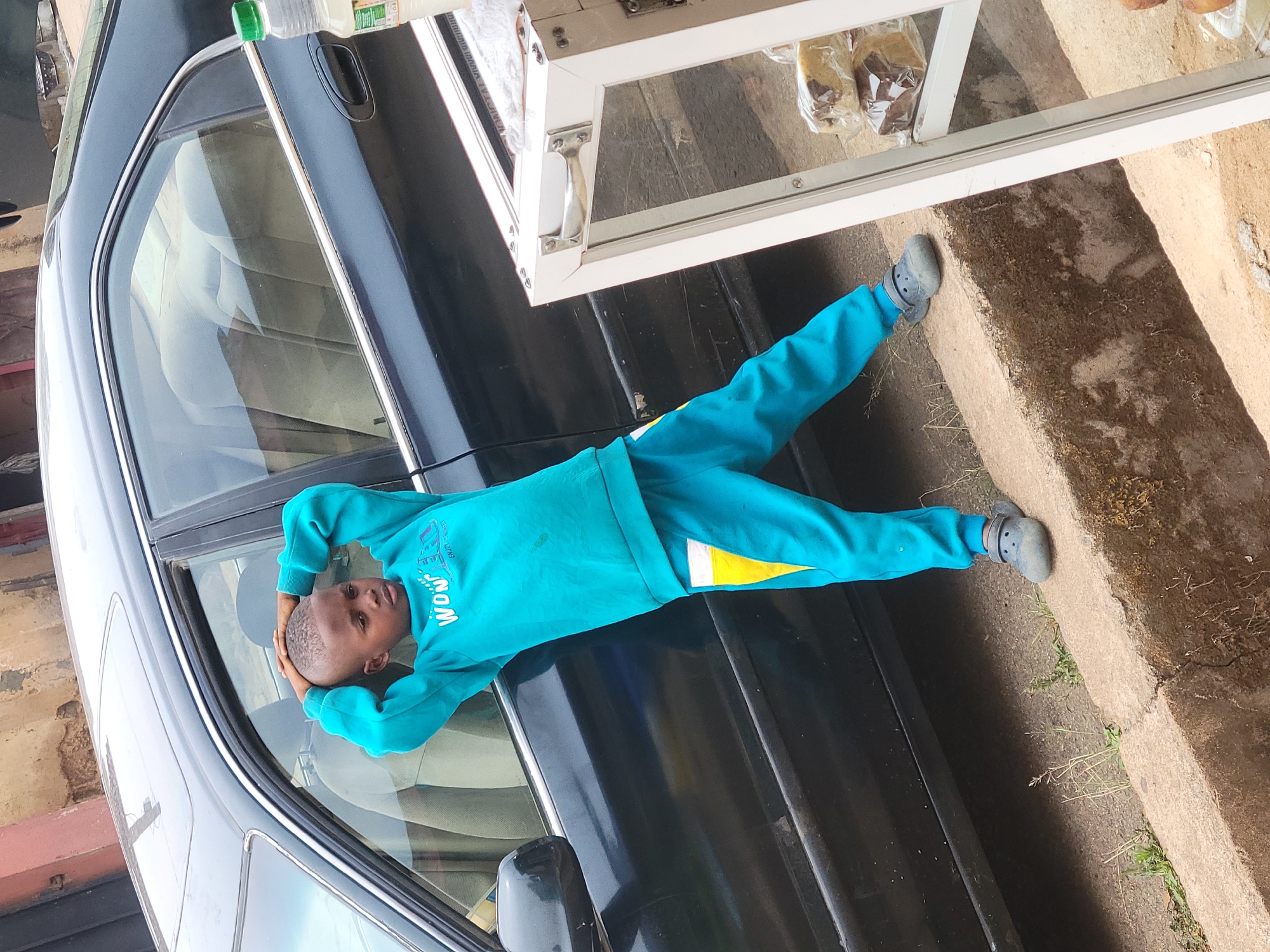A boy in a turquoise tracksuit and grey clogs lies on the hood of a black car next to a display case with pastries, likely in Jos, Nigeria. The boy has his hands behind his head. The car is parked next to a concrete ledge.