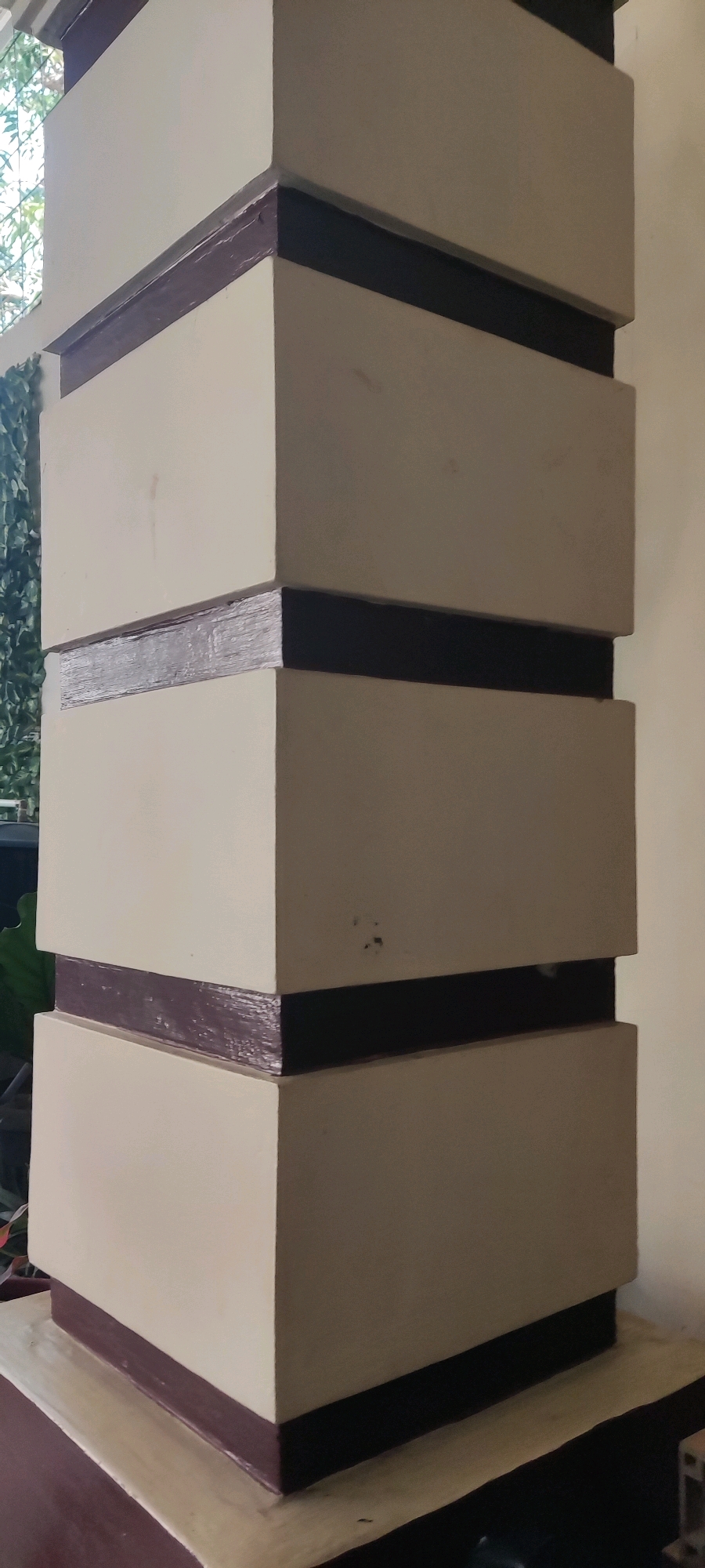 The image shows a close-up of a decorative pillar with a tiered design. The pillar is painted in a light beige color and features horizontal bands of dark brown and black. These bands are applied at intervals, creating a segmented appearance. The pillar has a slightly textured finish, and some minor imperfections are visible, such as small dark marks on the beige sections. The base of the pillar appears to rest on a platform that is also painted in similar colors. In the background, out of focus, there is greenery, suggesting an outdoor or semi-outdoor setting. The location is identified as Japura Kidul, Indonesia.
