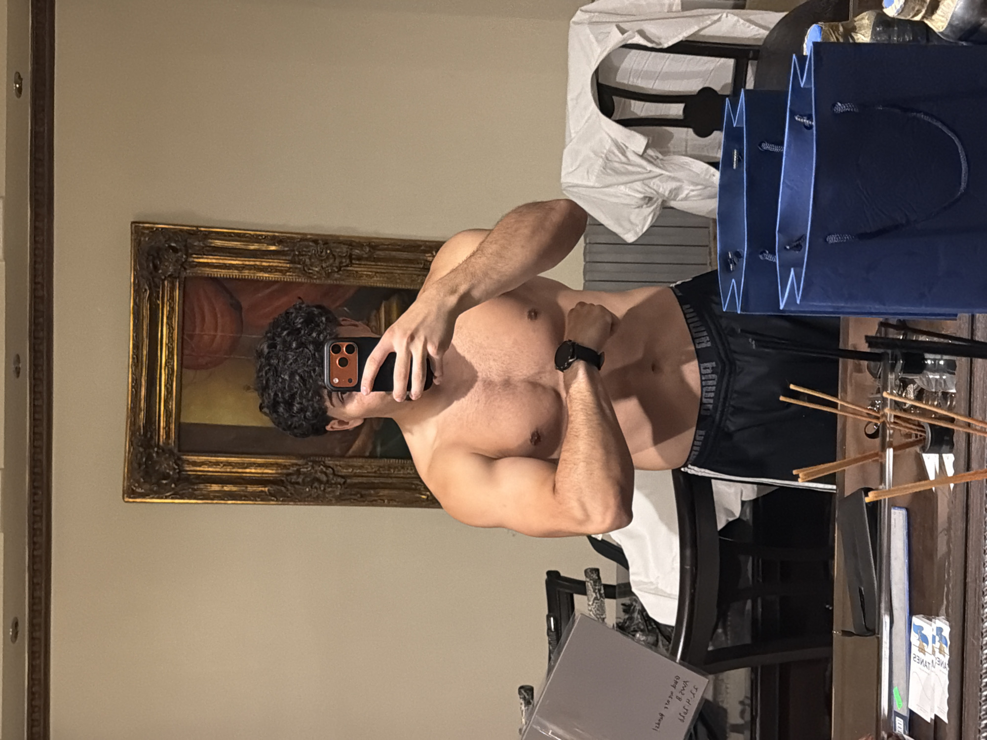 In a room with light beige walls and ornate crown molding, a shirtless individual is captured in a mirror selfie. The person has dark, curly hair and is holding a smartphone to take the picture. They are wearing black athletic shorts with a prominent logo waistband. The reflection shows their muscular build, with arms flexed. Behind them, a large, antique-style gold-framed painting hangs on the wall. To the left of the painting, a dark wooden chair is visible. In the foreground, a wooden surface reflects the scene. On this surface, there are several blue gift bags, a stapler, a clear plastic ruler with a green label, and other assorted items including what appear to be incense sticks. A white shirt is draped over the back of a chair in the background. The lighting suggests an indoor, possibly domestic setting, though no specific urban indicators place it in Sarouja Municipality, Syria.