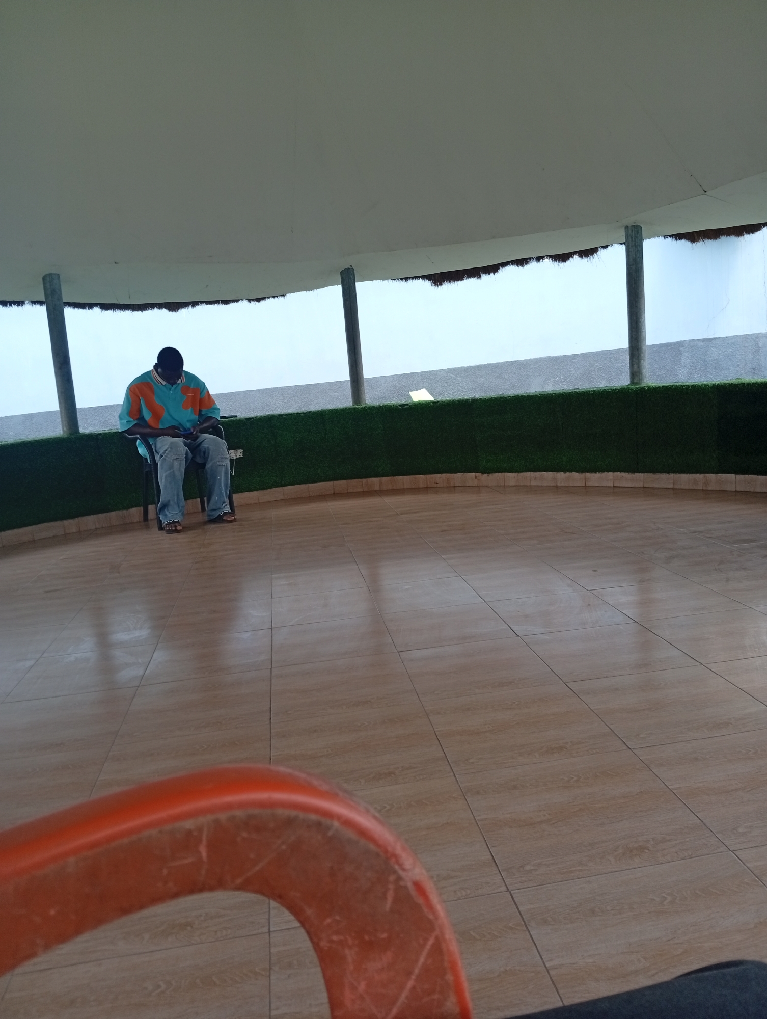 A person is seated on a chair in an open-air structure. The person wears a blue and orange patterned shirt and jeans. The floor is tiled. The structure features a white roof supported by pillars. Green artificial turf runs along the wall. The city is Adankolo, Nigeria.