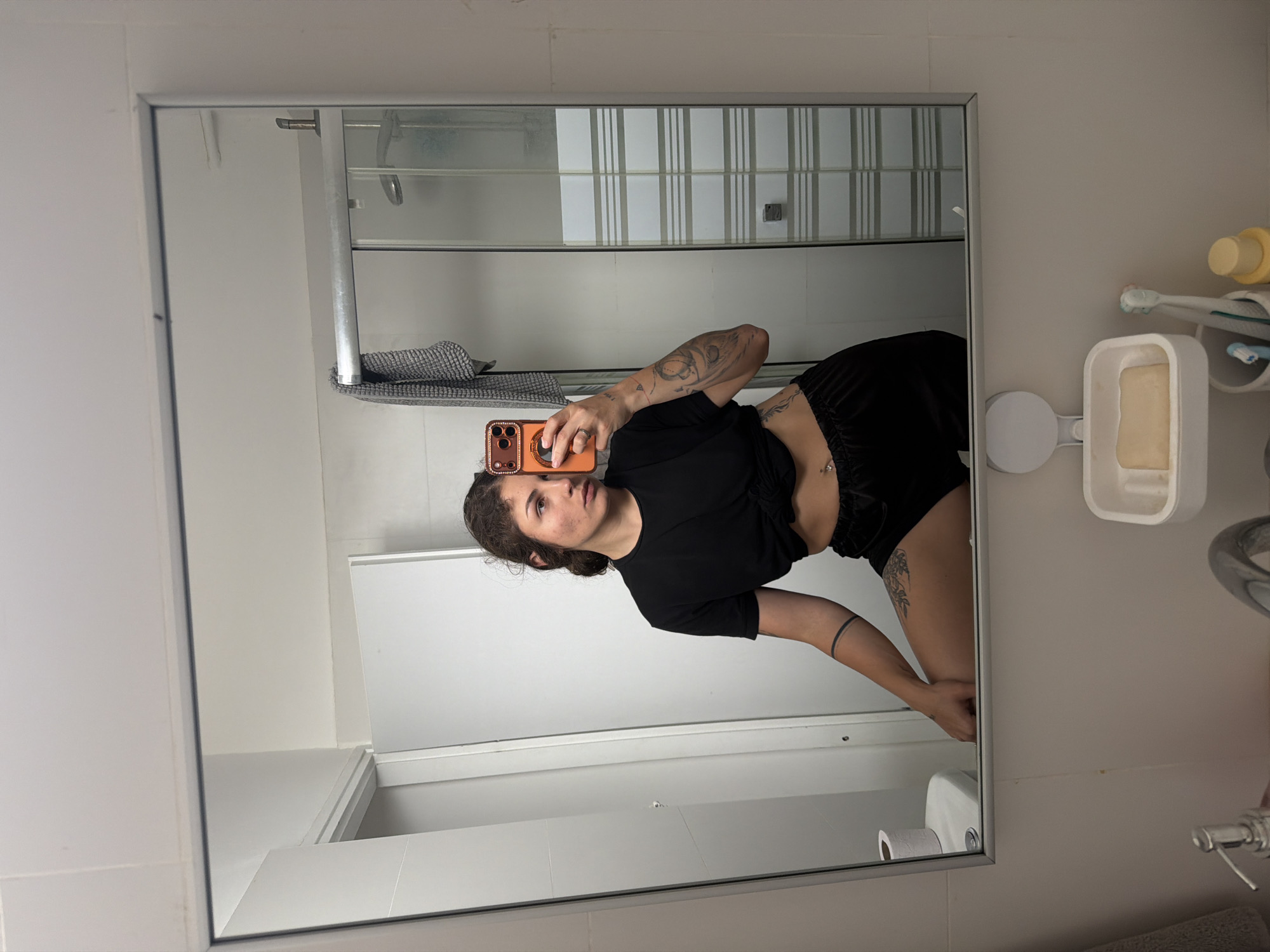 A mirror selfie taken in a bathroom. A person with brown hair and multiple tattoos on their arm is holding an orange phone and facing the mirror. The person is wearing a black crop top and black shorts. The bathroom features a shower with a gray towel, a white soap dish, a toothbrush holder, and a toilet paper roll. The location is Kiryat Bialik, Israel.