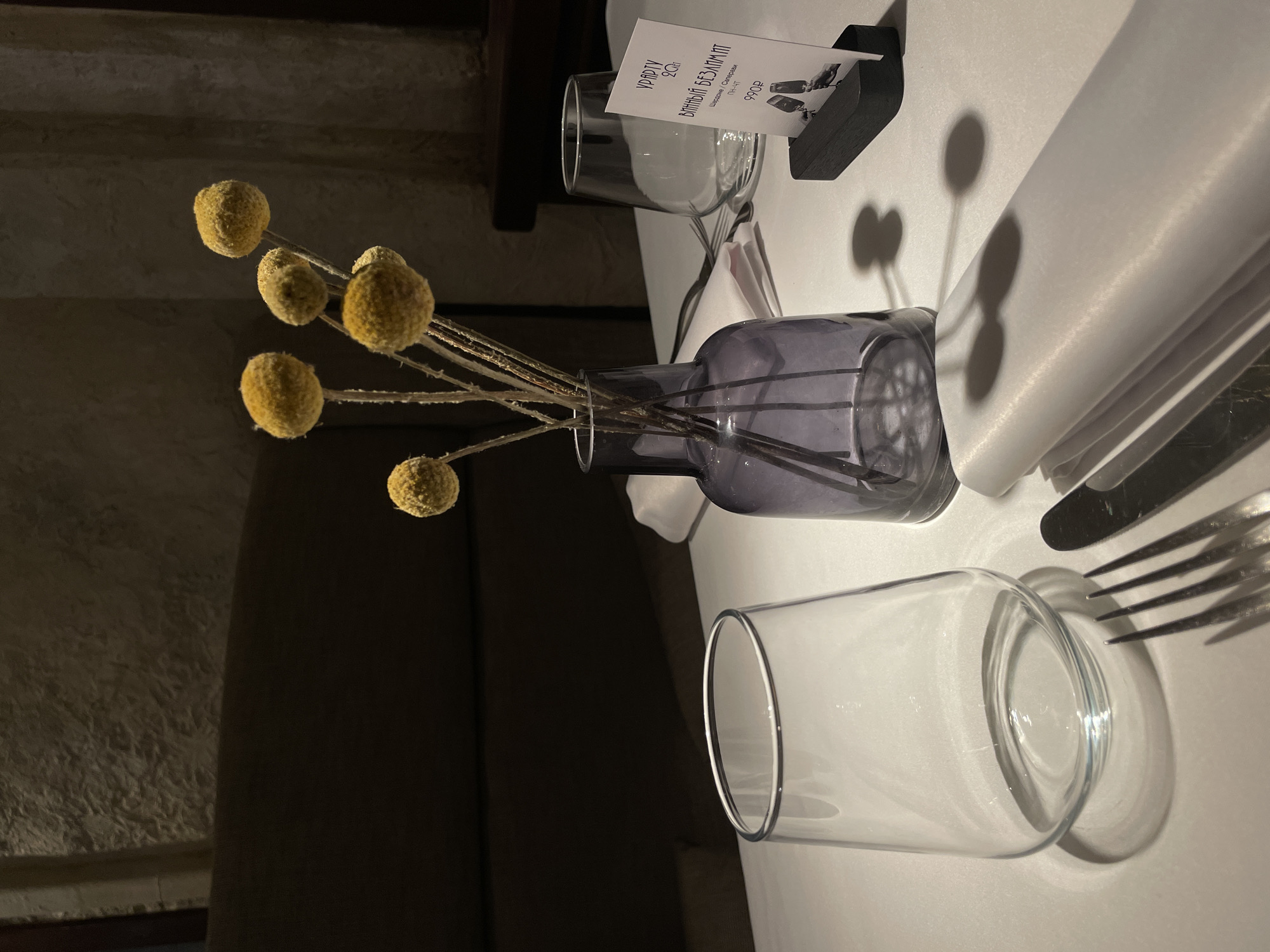 The image presents an intimately lit indoor table setting, likely within a restaurant or cafe in Krasnoyarsk, Russia. The focal point is a white tablecloth, meticulously arranged with various dining elements.
On the left, a dark purple glass vase contains several dried yellow spherical flowers, commonly known as 'billy buttons' or Craspedia. These flowers cast distinct, elongated shadows onto the bright white tablecloth. Adjacent to the vase, a neatly folded white napkin is positioned beside a fork and knife. A clear, empty glass tumbler rests further along the table.
In the background, a menu or information card stands upright in a black holder. The card displays text in Cyrillic script. Prominently featured is "ВИННЫЙ БЕЗЛИМИТ," translating to "WINE UNLIMITED." Below this, "ШАМПАНЬ | СТОЛОВЫЙ" ("Champagne | Table wine") is visible, alongside a price of "990₽" (990 Russian Rubles), indicating a special offer. A small graphic of wine glasses is also printed on the card. Another line, "УРГАРТУ 20ч.", is present, which could refer to a brand name, time duration, or portion detail, though its exact meaning is not fully clear without additional context.
The lighting is dim and ambient, creating a sophisticated and calm atmosphere. No people are visible, and the table appears set and ready for diners, highlighting the prepared and aesthetic environment of the establishment.