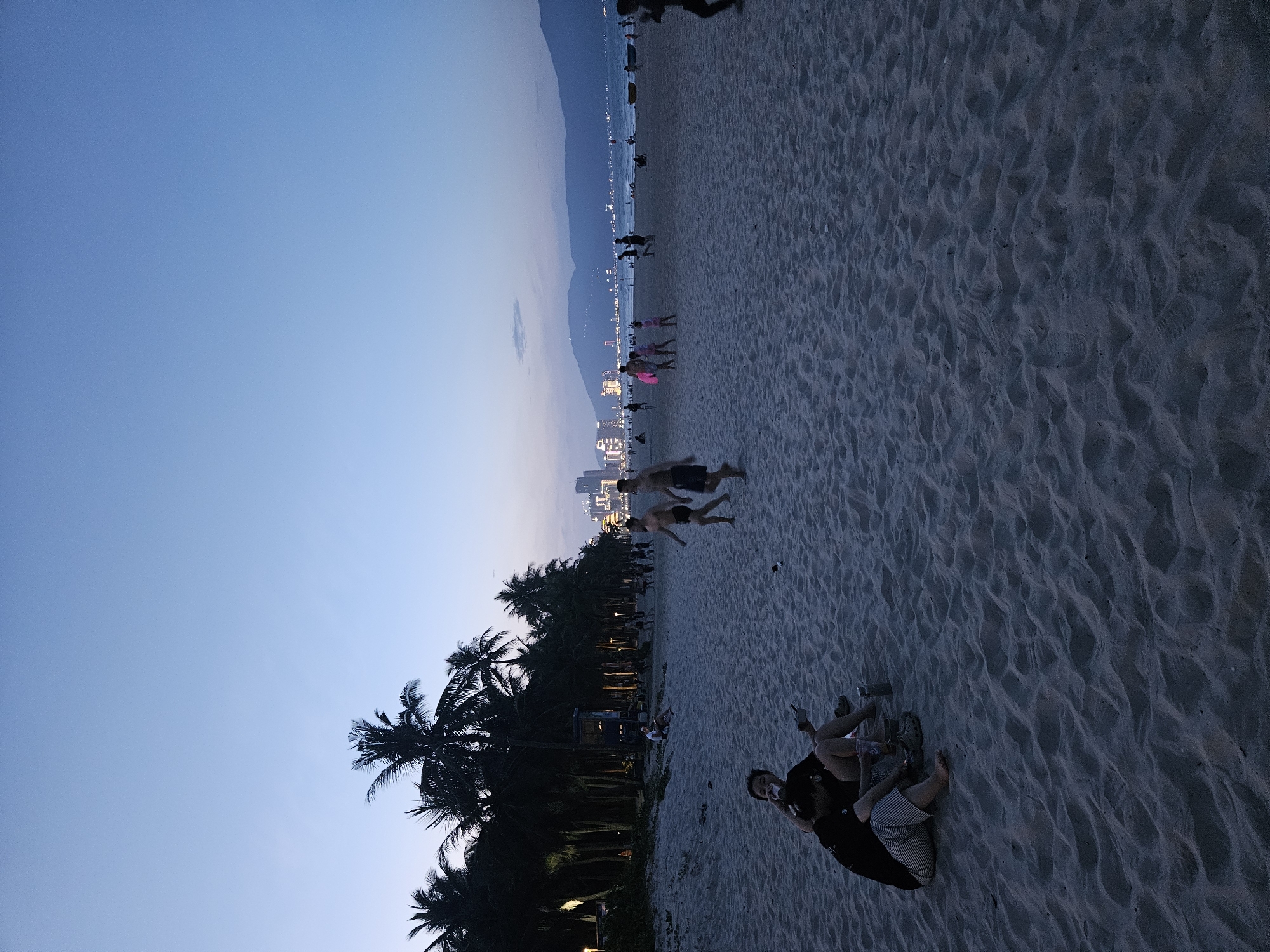The image depicts a beach scene in Da Nang, Vietnam, during dusk. The sky is a gradient of deep blue, transitioning to a lighter shade near the horizon, suggesting the sun has recently set. The beach is sandy, with visible footprints and ripples in the sand.
Several individuals are scattered across the beach. In the foreground, two people are sitting on the sand, one of them looking towards the camera and holding a phone. Further down the beach, a group of people are visible, some walking and others appearing to be playing. In the middle ground, a couple of individuals are seen in mid-air, possibly jumping or engaged in some playful activity.
Along the coastline, a line of palm trees forms a silhouette against the dusky sky. Beyond the trees, city lights twinkle, indicating a developed urban area. The ocean is a dark expanse, with subtle reflections of the lights. The overall mood of the image is relaxed and serene, characteristic of a beach at the end of the day. The weather appears to be clear. No visible text is present in the image.